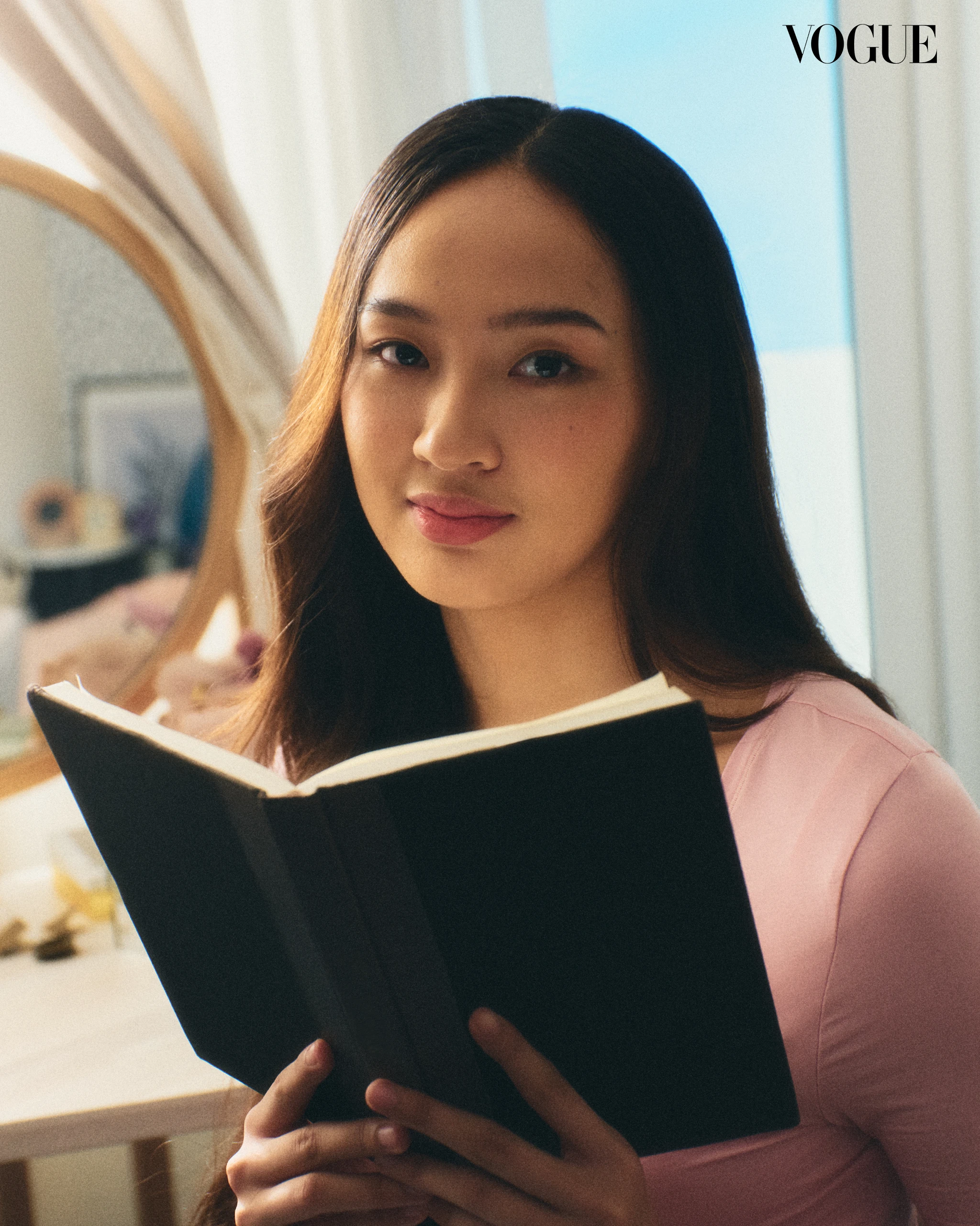 Woman in a pink top reading a black notebook indoors; Vogue logo in the corner.