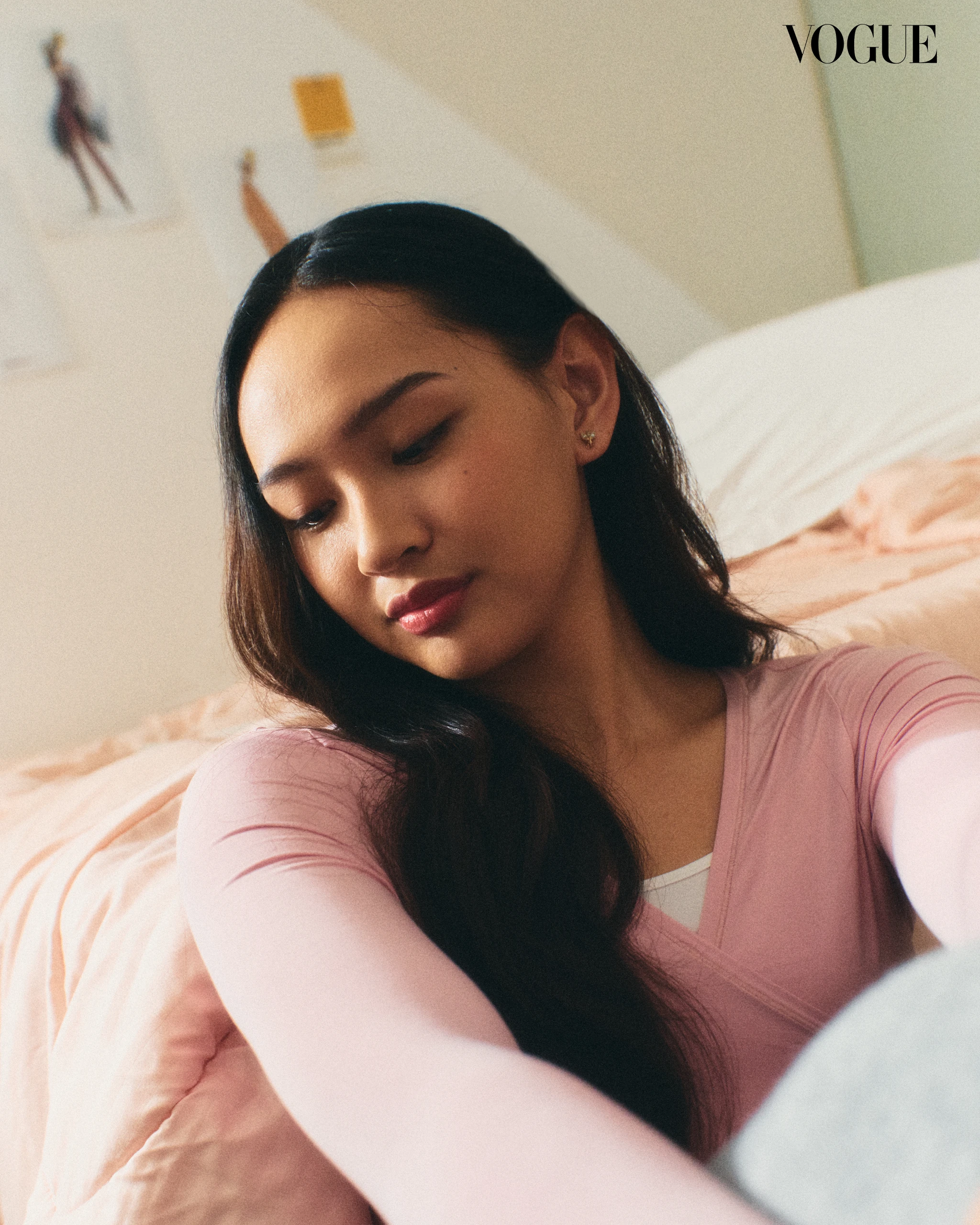Close-up selfie of a woman with long dark hair wearing a pink top, resting on a peach bedspread with a Vogue logo in the top-right corner.