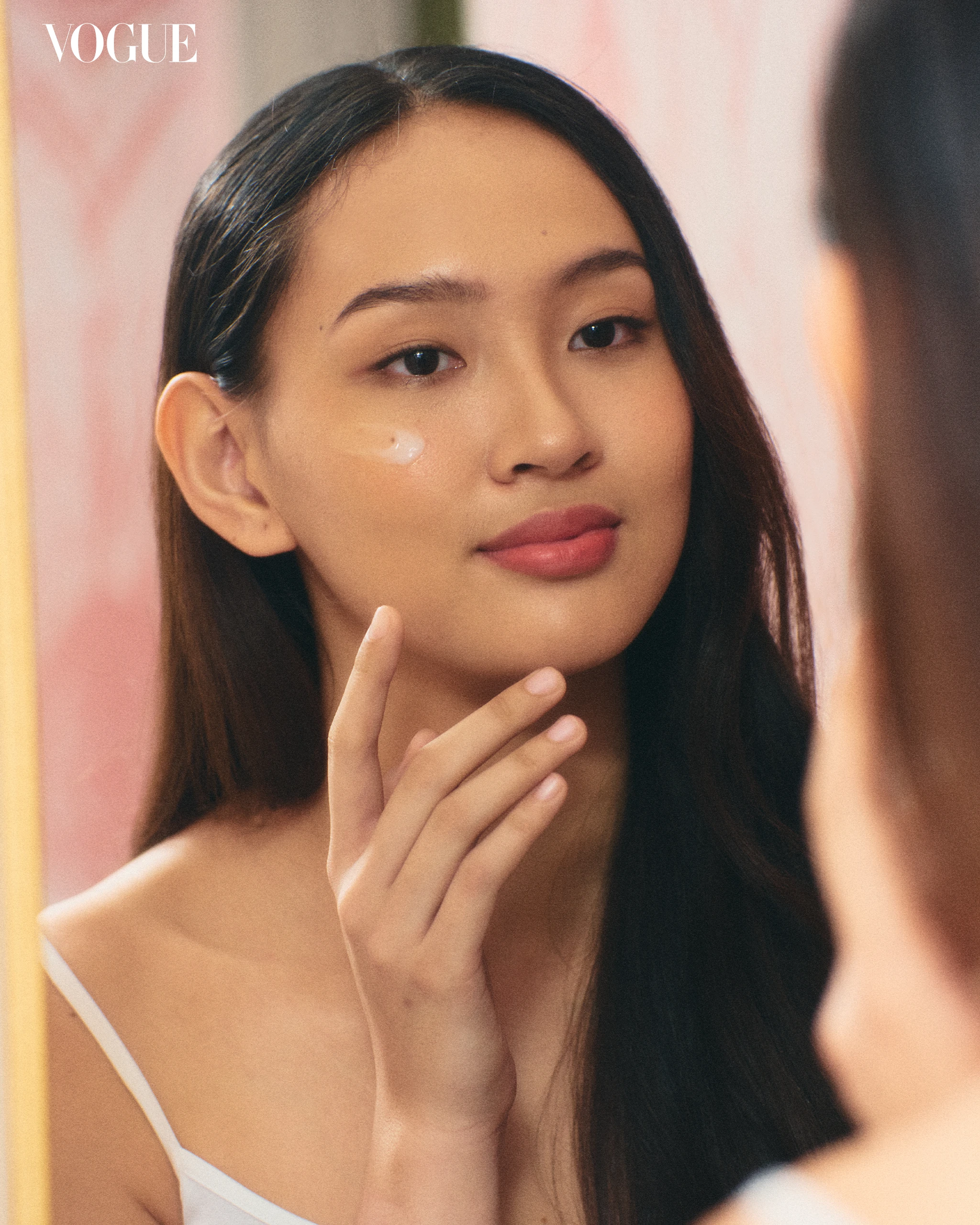 Close-up of a woman applying moisturizer under her eye, pink background, Vogue logo in the corner.