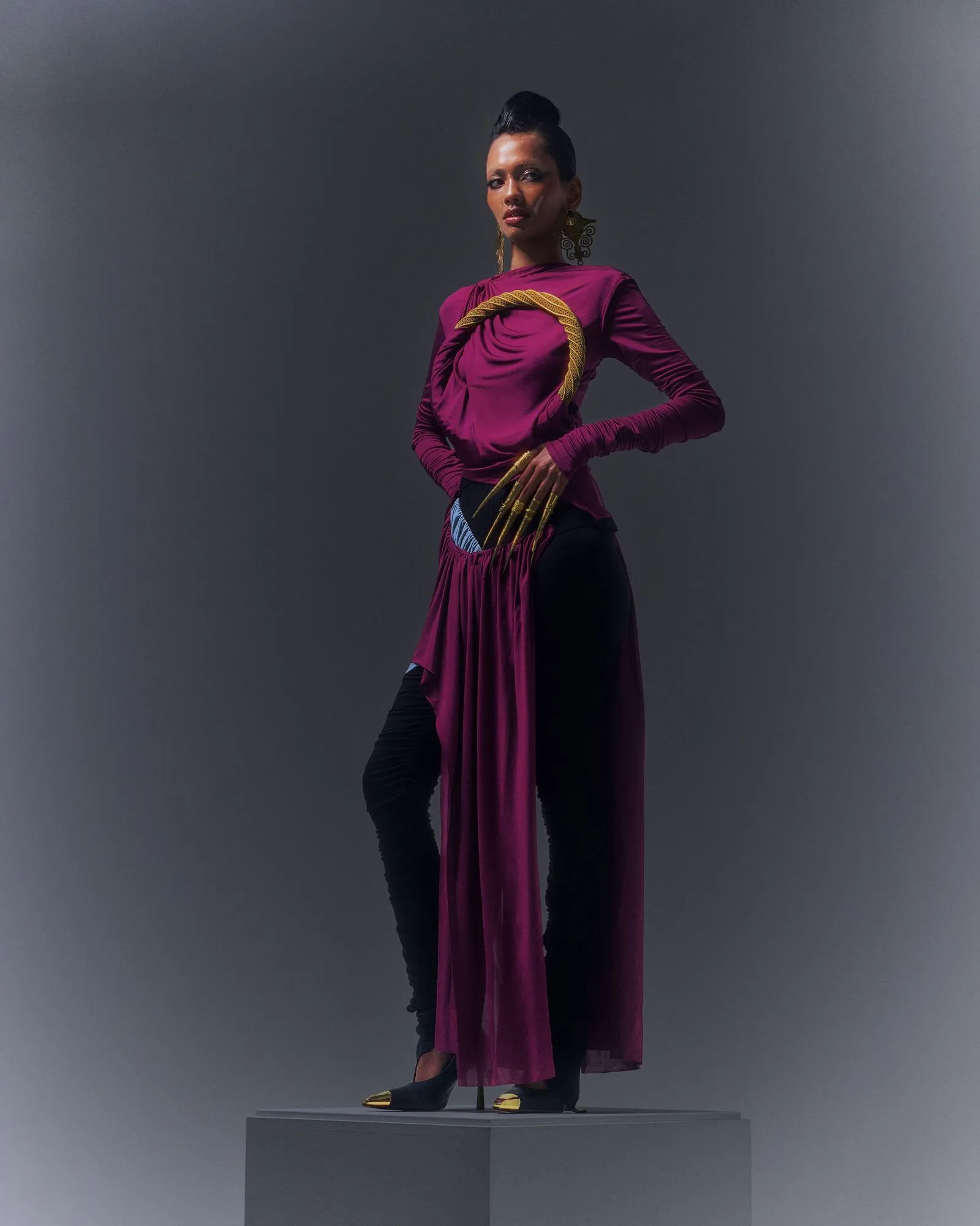 Model in a magenta draped gown with gold jewelry, standing on a pedestal against a gray backdrop.