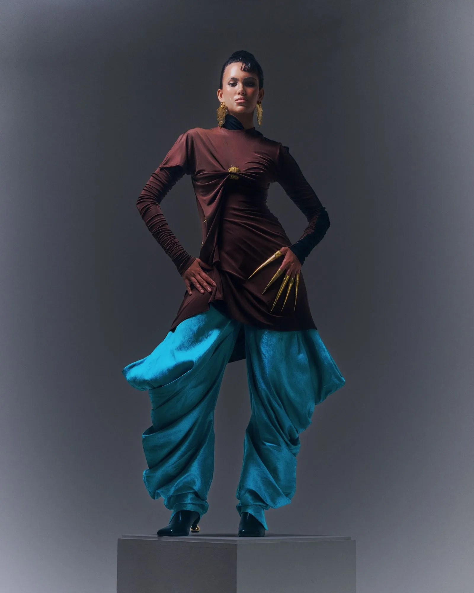 Fashion model standing on a pedestal, wearing a burgundy long-sleeve top with teal billowy pants and large gold earrings.