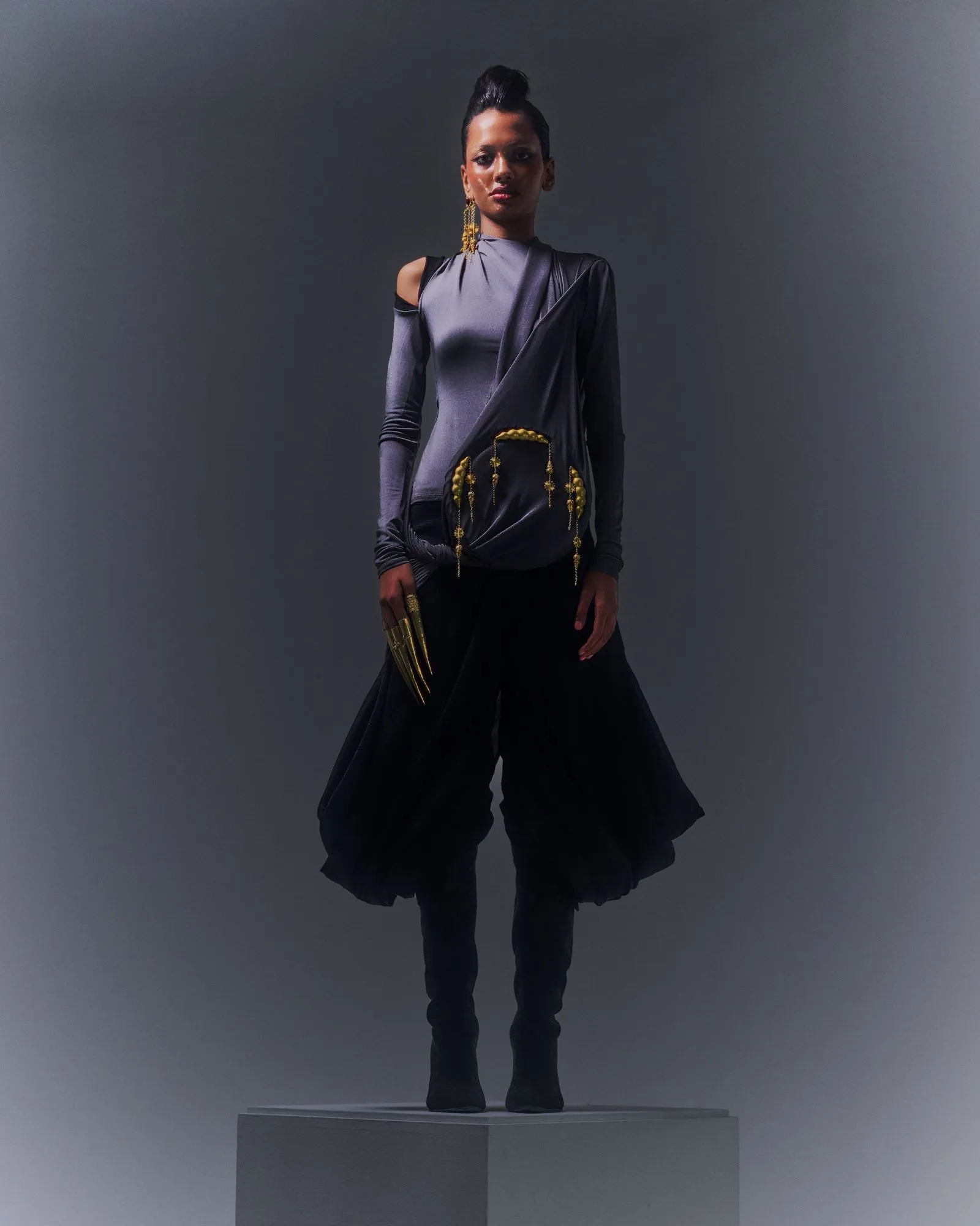 Model stands on a platform wearing a metallic gray draped top with cut-out shoulders, black wide-leg pants, and gold jewelry against a dark backdrop.