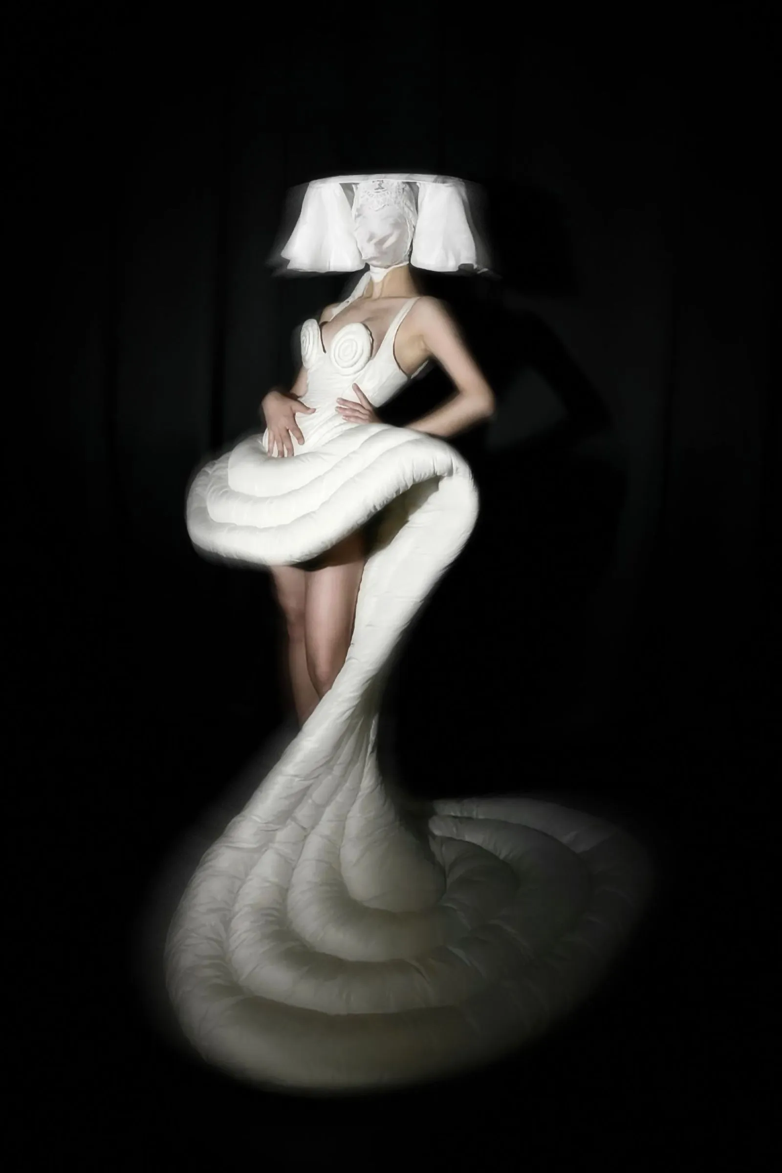 Model wearing a sculptural white gown with spiral bodice and large ruffled skirt against a dark stage backdrop.