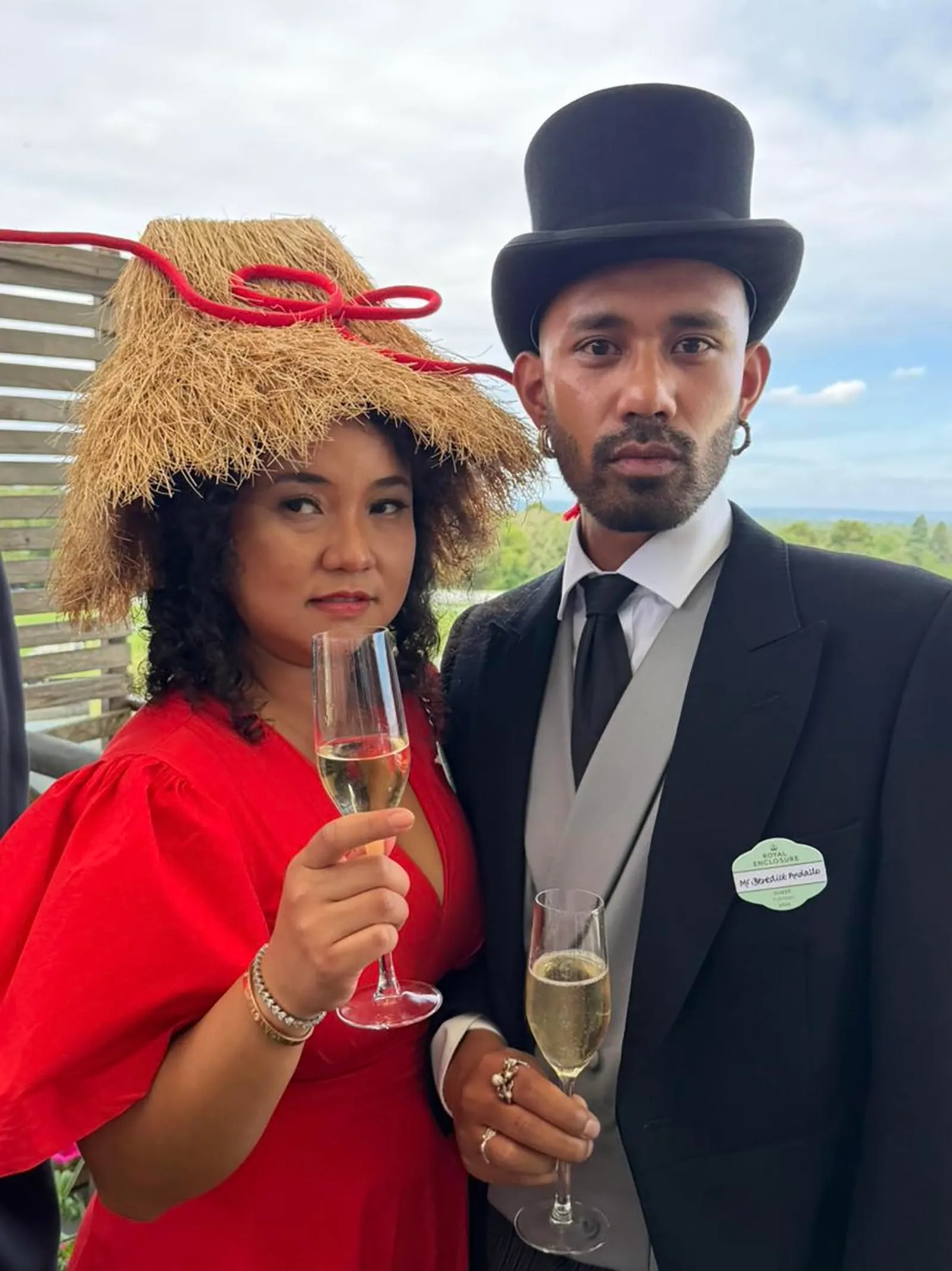 Two people posing in festive outfits, each holding a champagne flute; woman in red dress with a straw hat, man in a suit with a top hat outdoors.