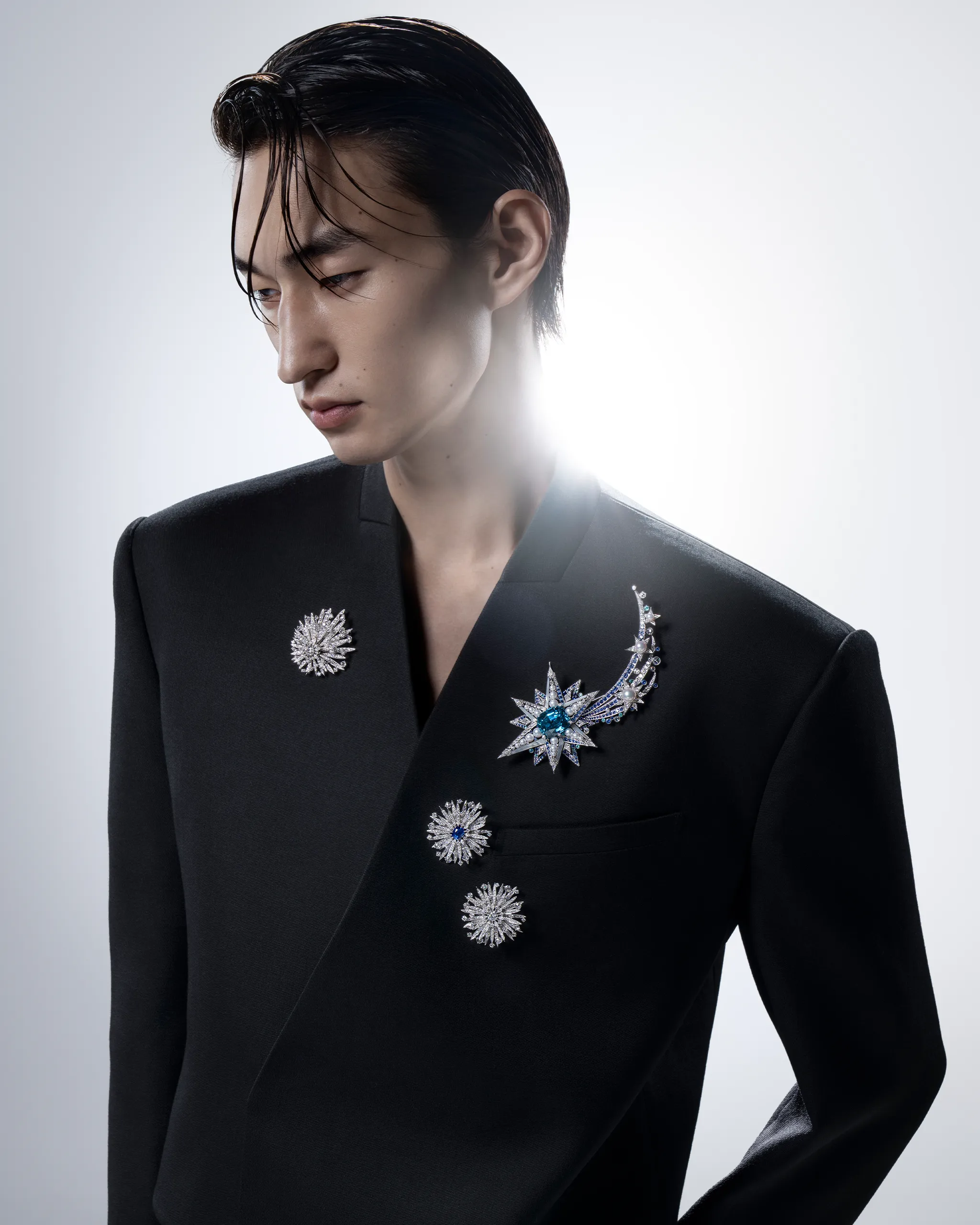Person wearing a black blazer adorned with large jeweled brooches on the lapel and chest, slicked dark hair, looking downward to the left.