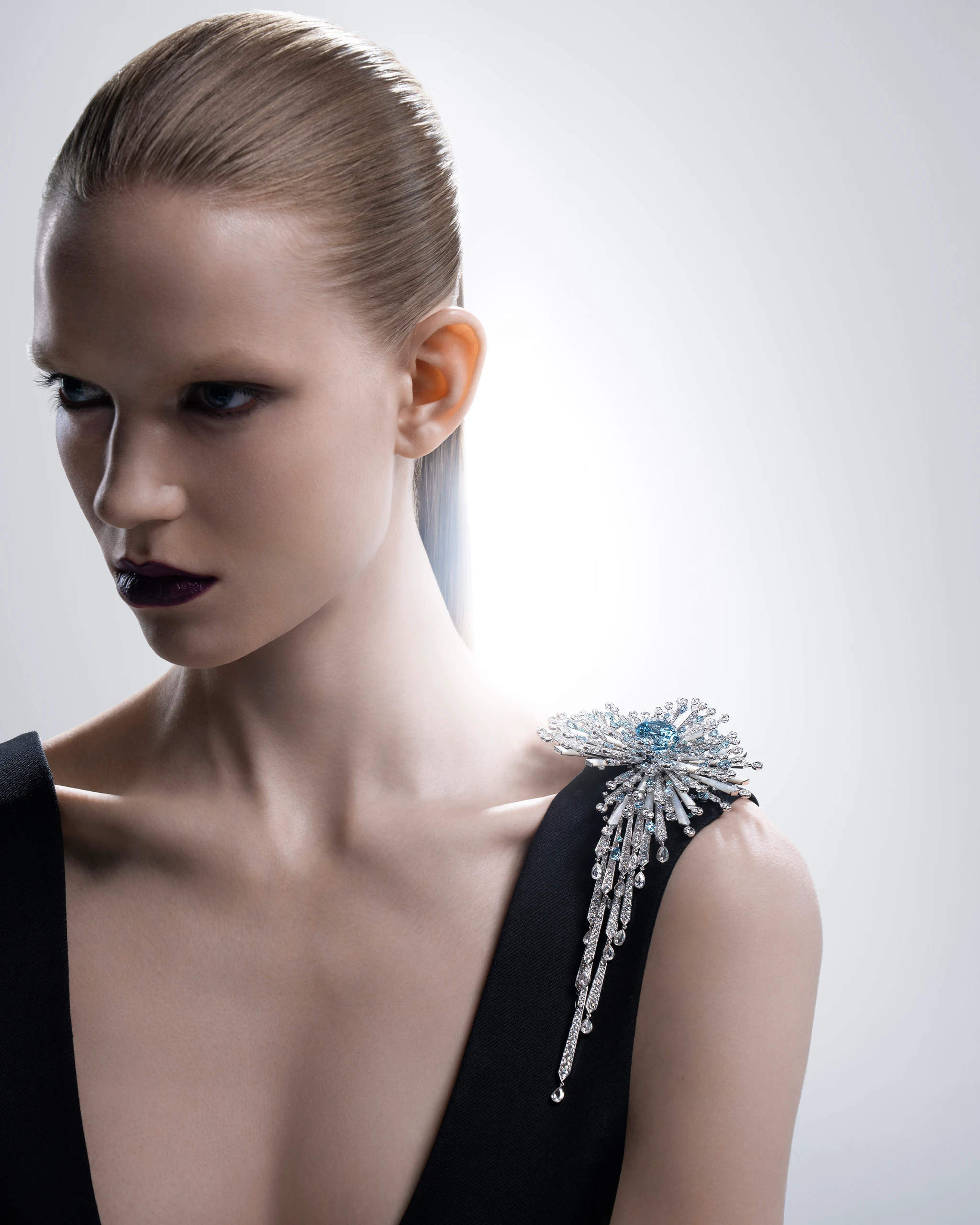 Portrait of a woman in a black dress with a sparkling jeweled shoulder ornament, facing left against a light gray backdrop.