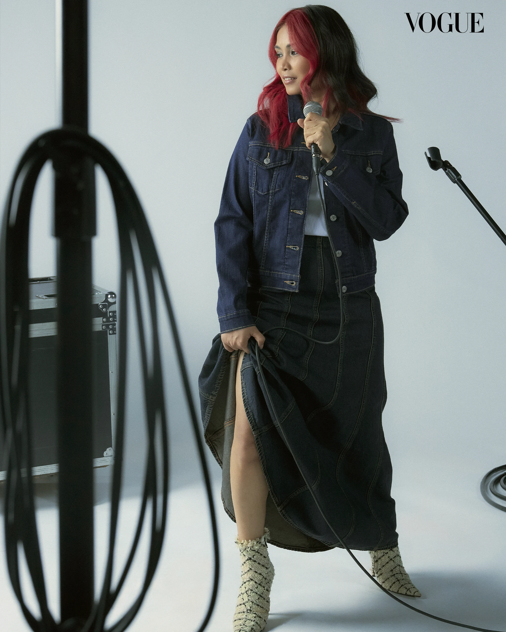 Female model with pink-streaked hair in a denim jacket speaks into a microphone in a studio with Vogue logo in the corner.