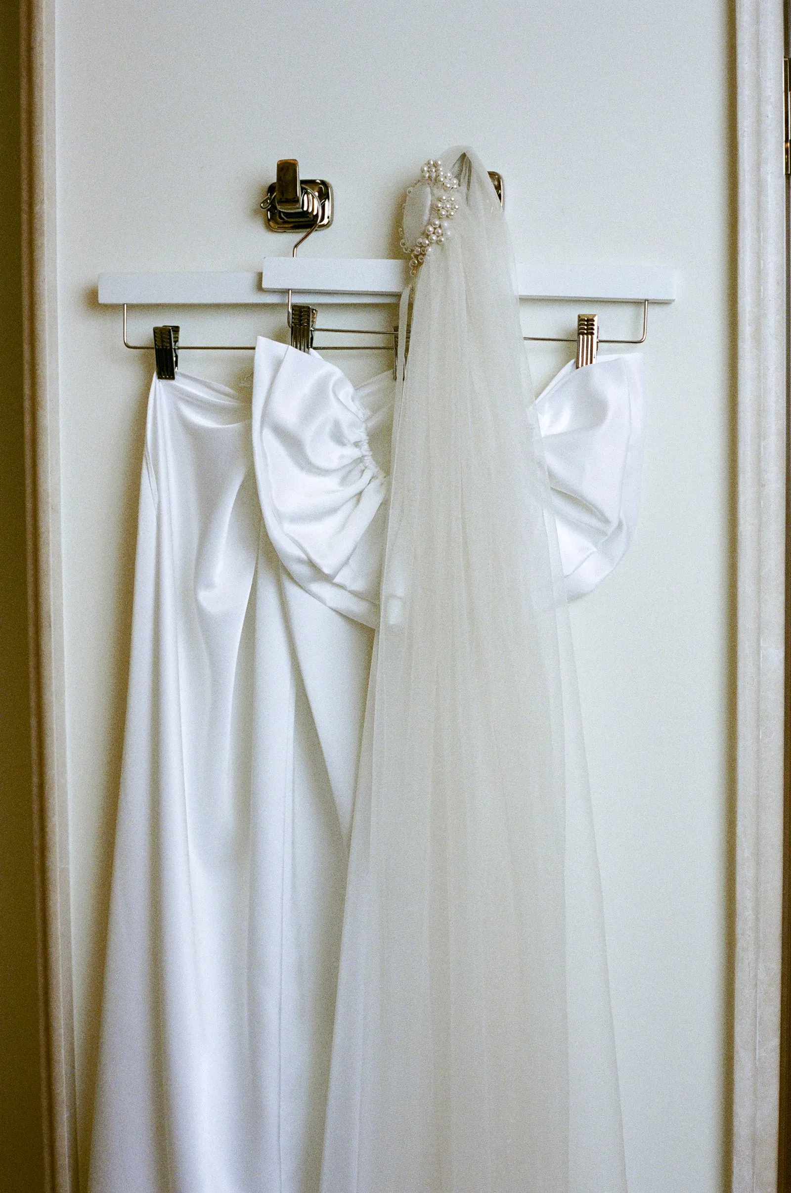 Wedding dress with veil and pearl accessory hanging on a metal hanger on a white door surface.