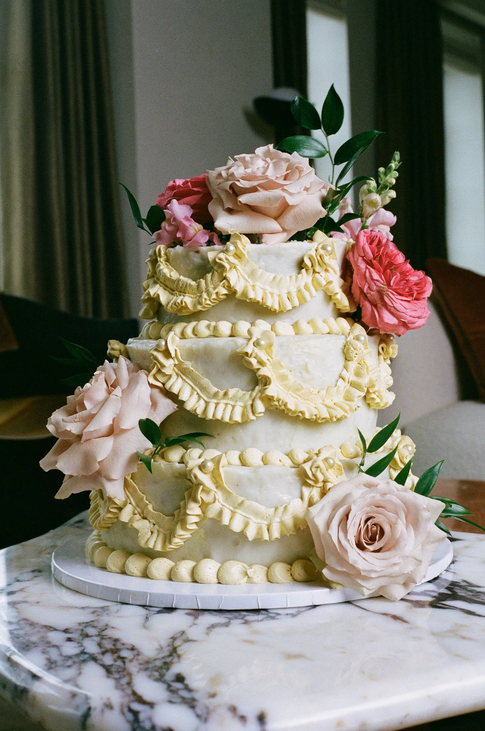 Multi-tier wedding cake with pale yellow ruffle trim and pink roses on top, set on a marble surface.