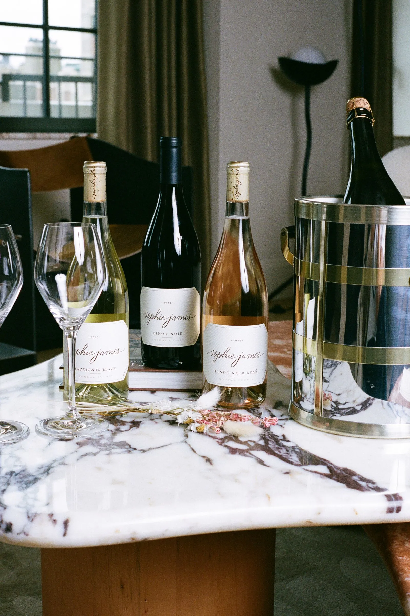 Three wine bottles (two white, one rosé) standing on a marble table next to a metal champagne bucket and two empty wine glasses.
