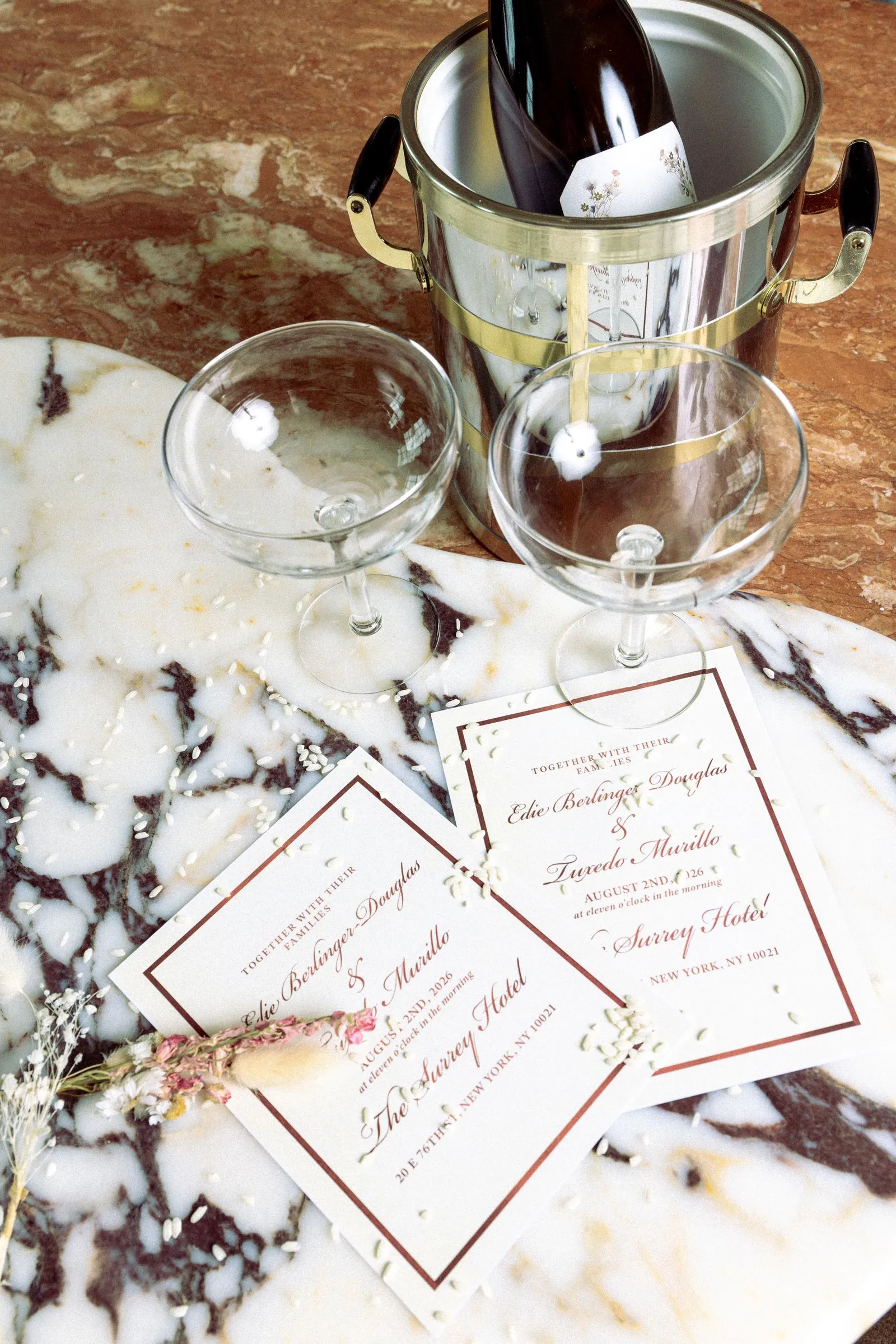 Champagne bottle chilling in a silver ice bucket with two empty wine glasses on a marble table and wedding invitations nearby.]','Champagne in an ice bucket with two stemmed glasses and wedding invitations spread on a marble surface.