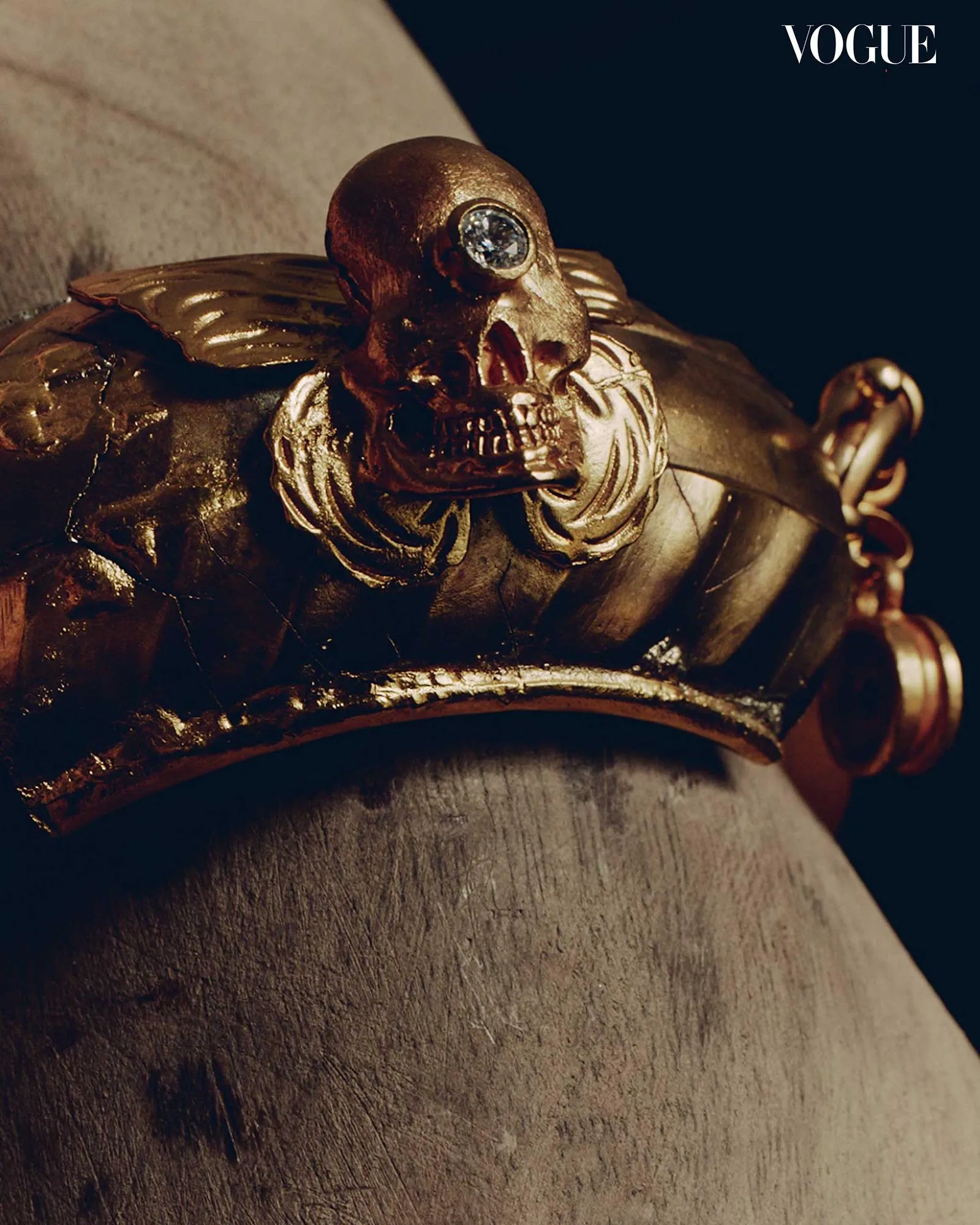 Close-up of a gold crown with a jeweled eye resting on a carved sculpture; dark background and a Vogue logo in the corner.