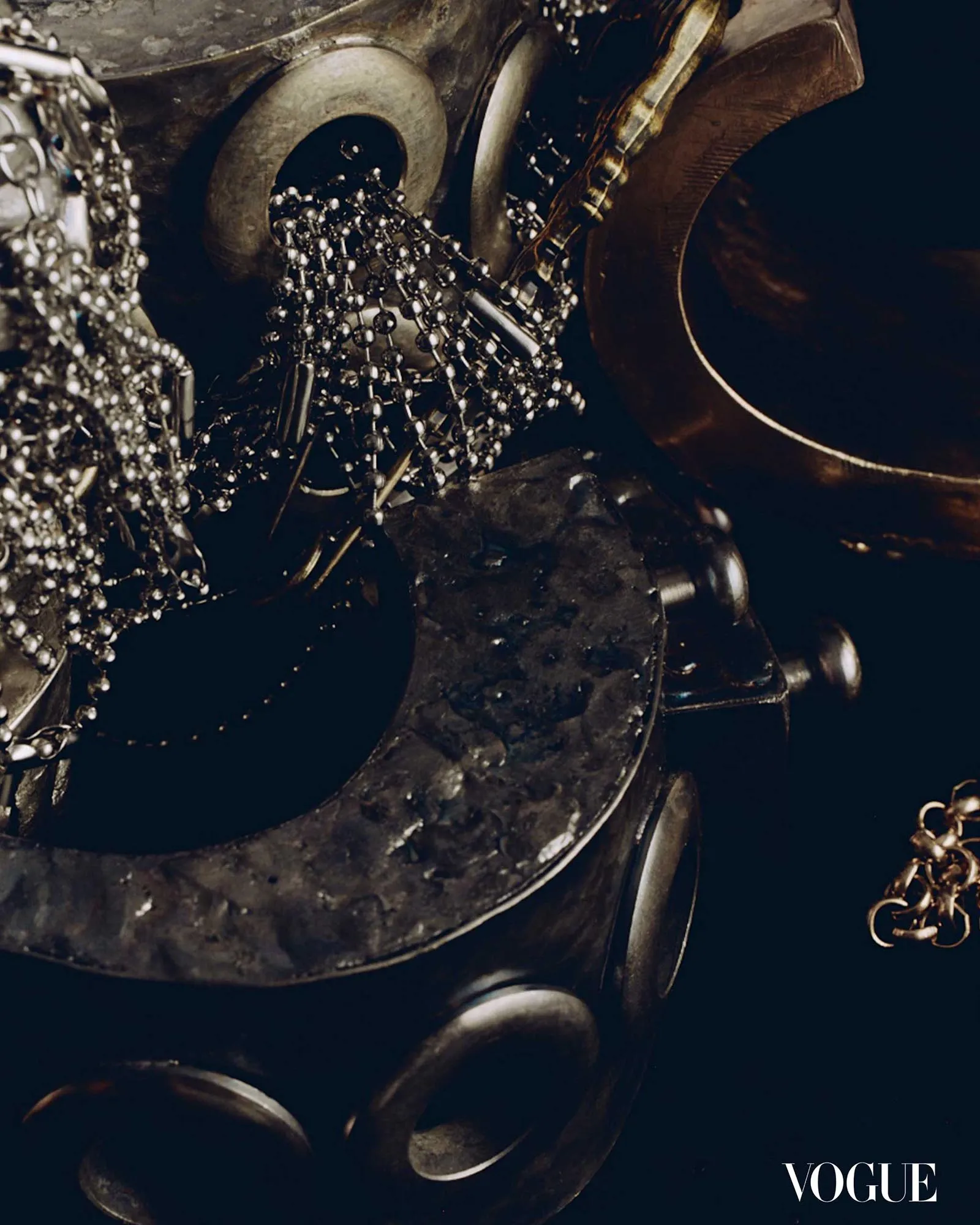 Close-up of metallic chains, beads, and dark metal surfaces on a textured fashion setup with Vogue logo in the corner.