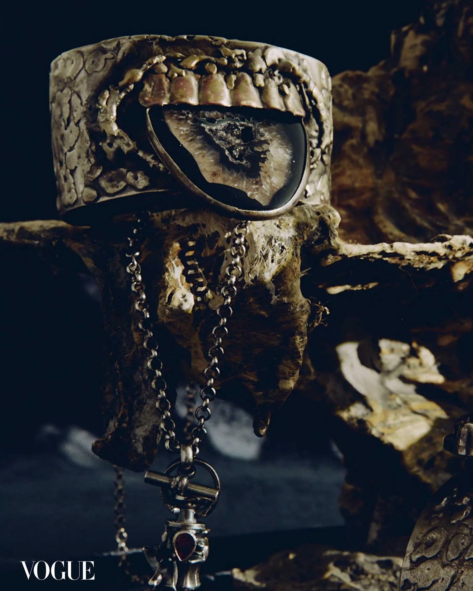 Close-up of ornate metal jewelry (chains, rings, pendant) draped over a textured sculpted bust, with Vogue in the corner.