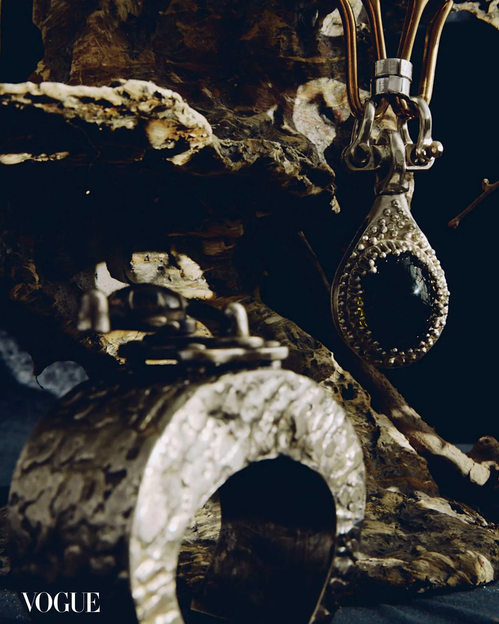 Close-up of an ornate metallic pendant with beaded edge hanging from rough rock in a cave setting, Vogue watermark visible in the corner