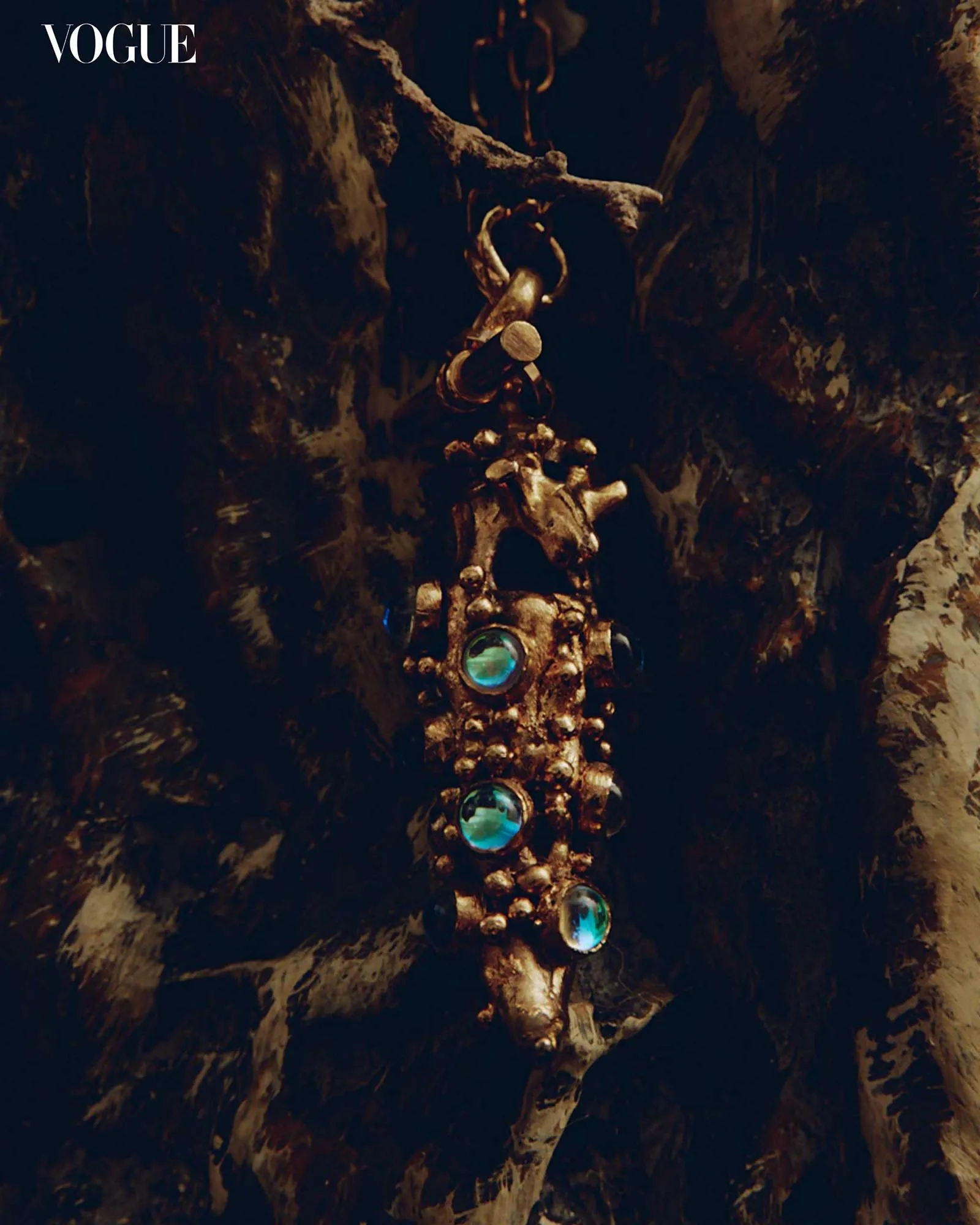 Close-up of a gold-toned necklace with blue gems displayed on a dark, rocky surface with Vogue in the upper-left corner.