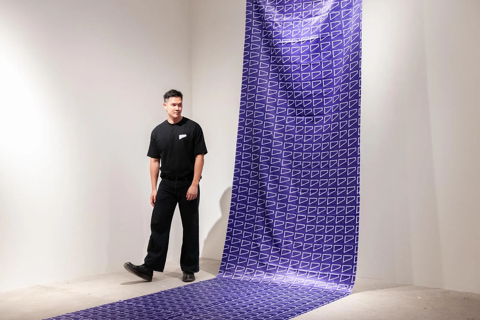 Man in a black shirt and dark jeans stands in a white gallery beside a tall purple fabric with white triangular pattern that drapes from ceiling to floor.