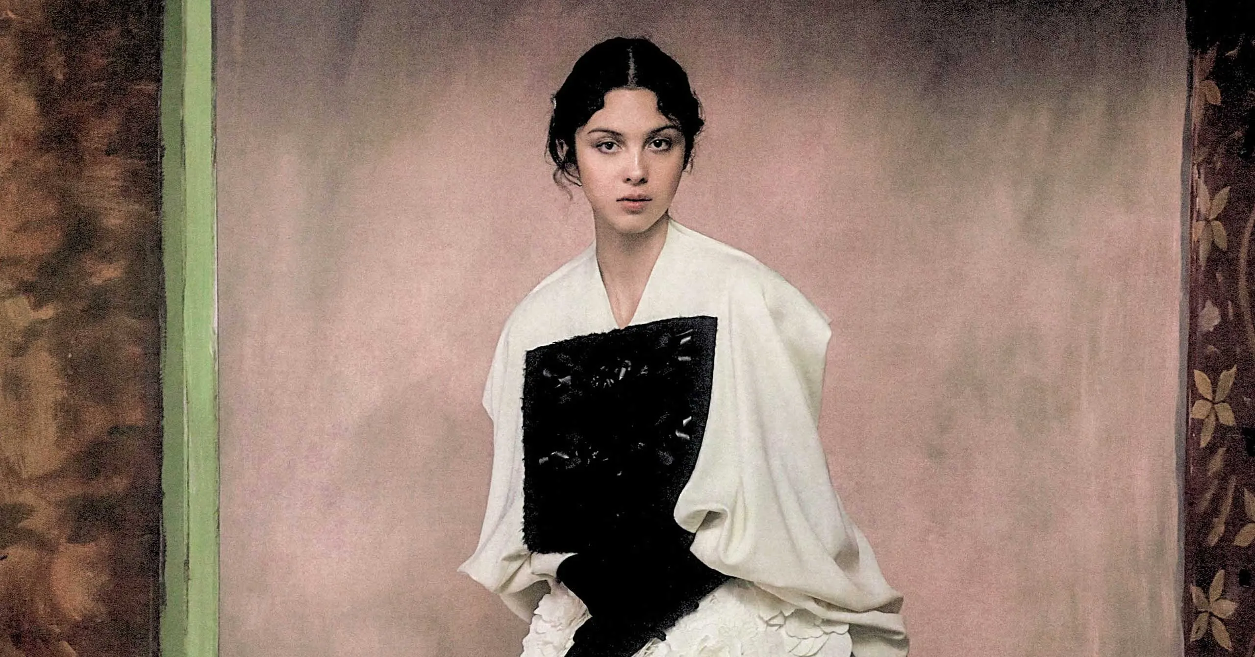 Portrait of a woman wearing a white robe with a black rectangular panel, standing against a pale pink backdrop.