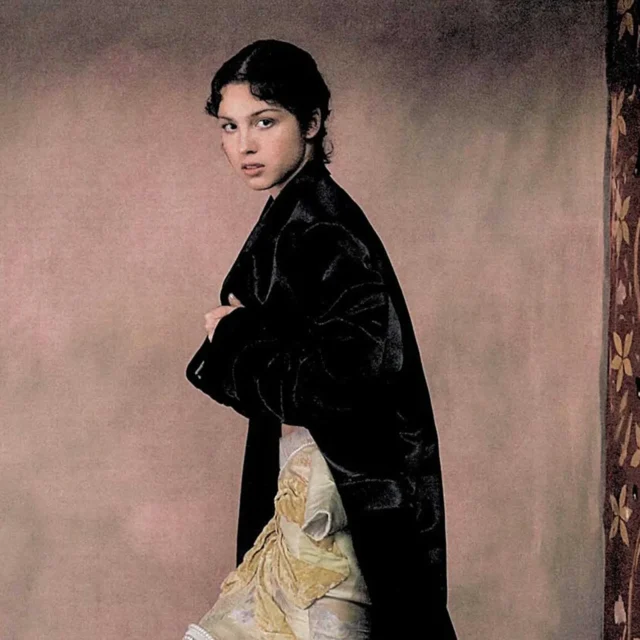 Side-profile of a woman wearing a black velvet gown, facing left against a muted pink backdrop.