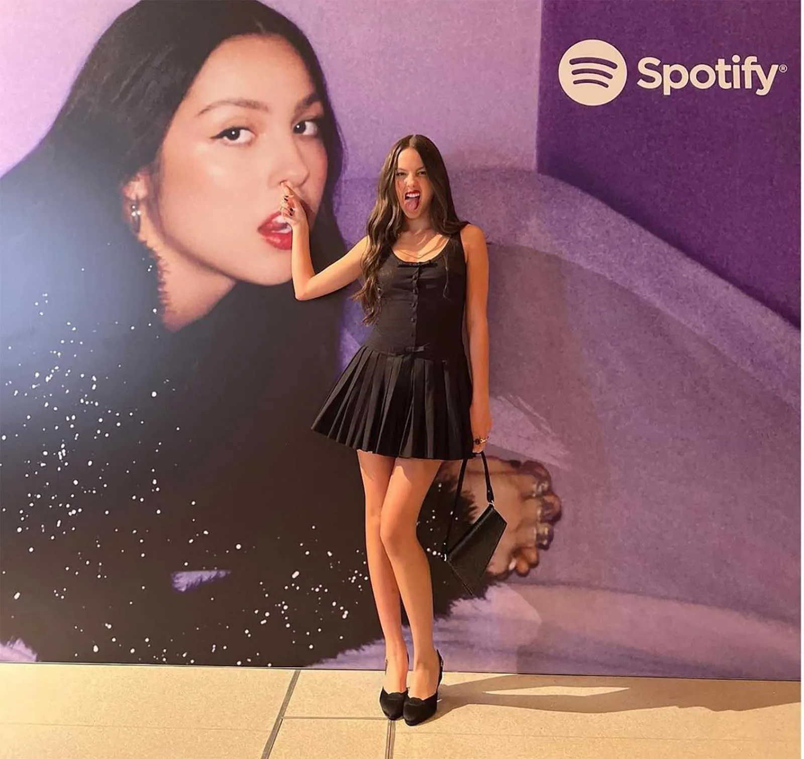 Woman in a black sleeveless dress posing with her finger near her nose in front of a purple Spotify backdrop.