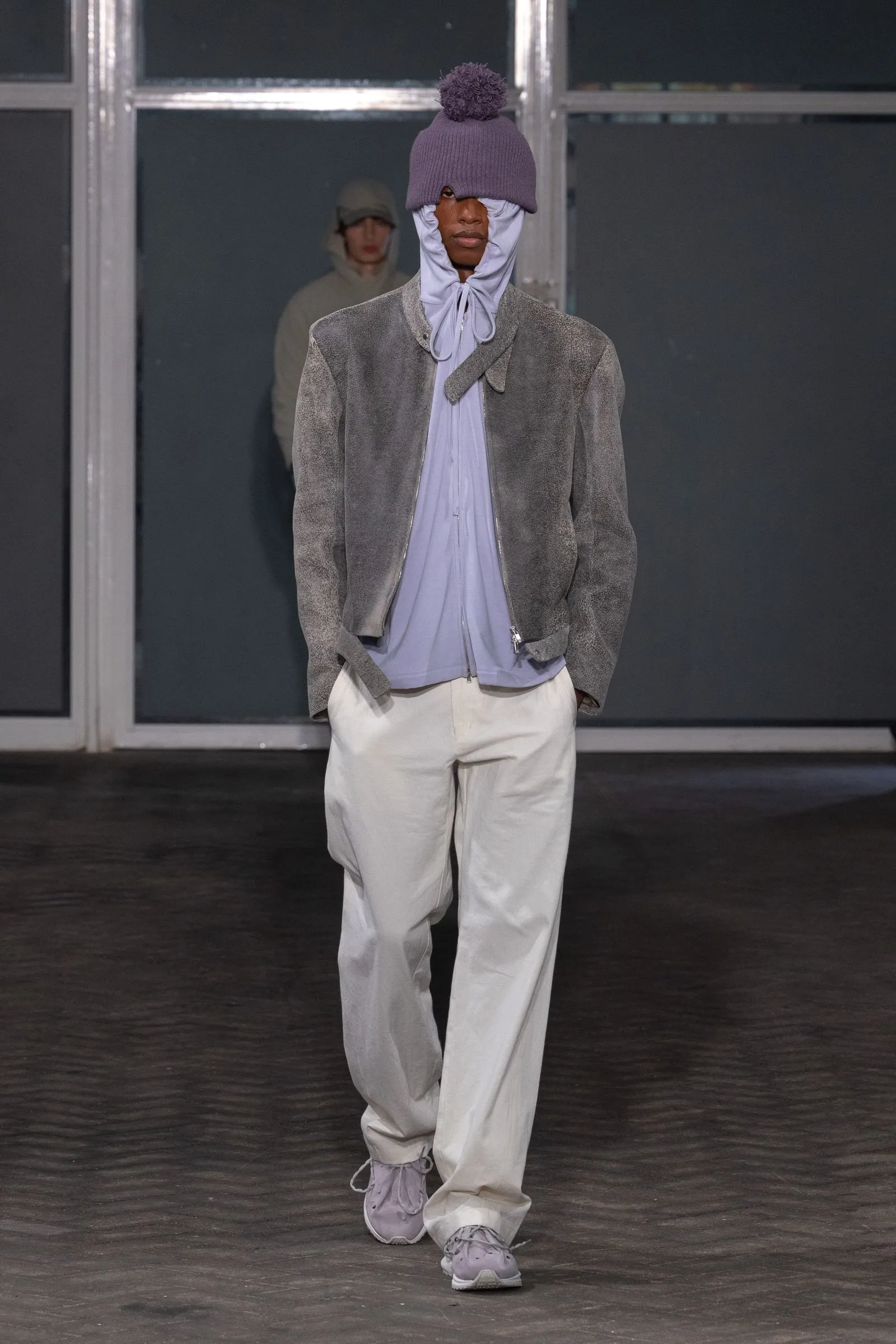 Runway model wearing a lavender hooded top under a gray suede jacket with light pants and a purple pom beanie.