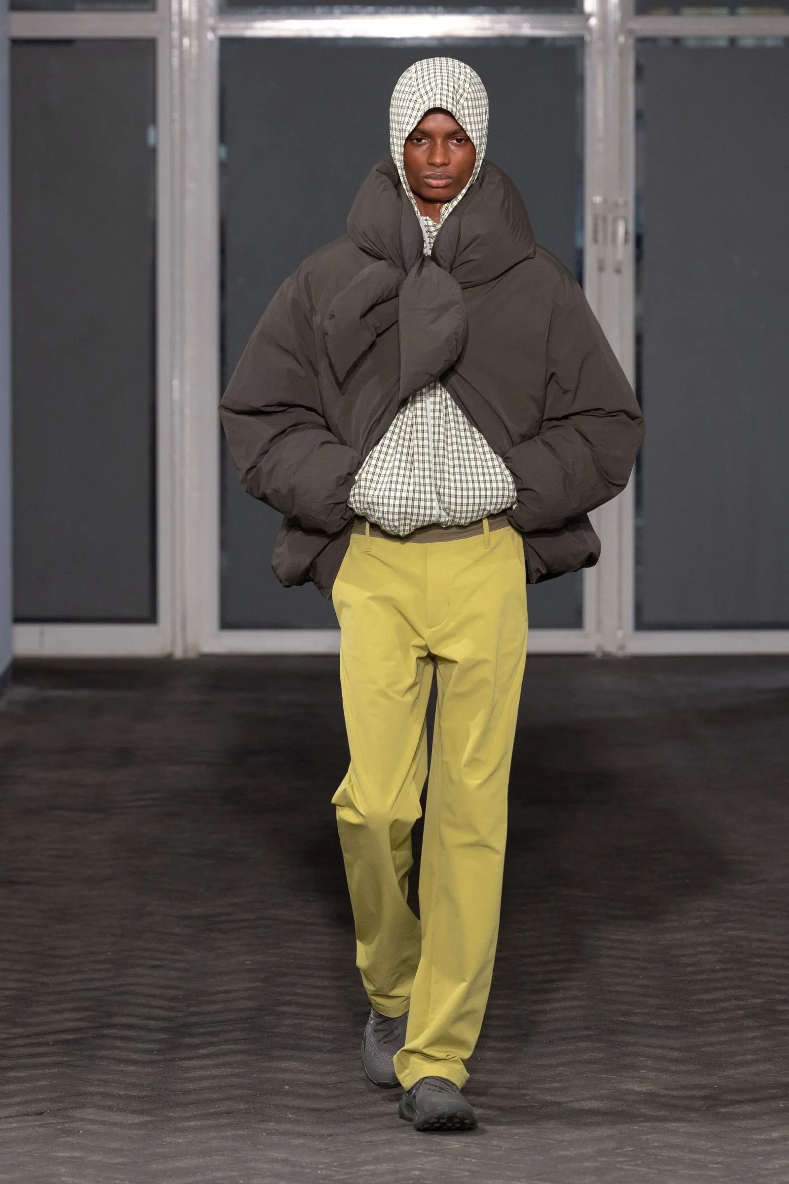Model on a runway wearing an oversized dark brown puffer jacket, plaid shirt, and bright yellow pants, with a checkered headscarf.