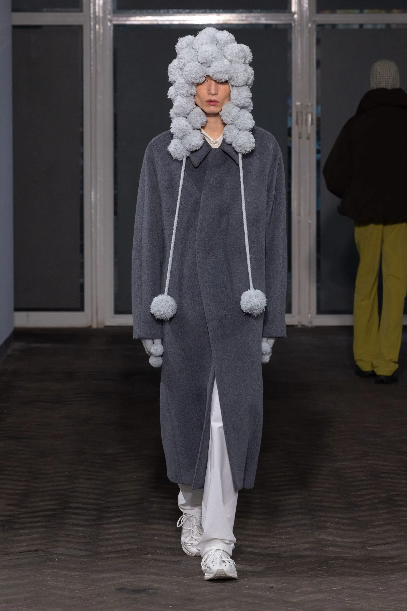 Model on a runway wearing an oversized grey coat with long white drawstrings ending in pom-poms and a fluffy grey pom-pom headpiece, white pants and sneakers.