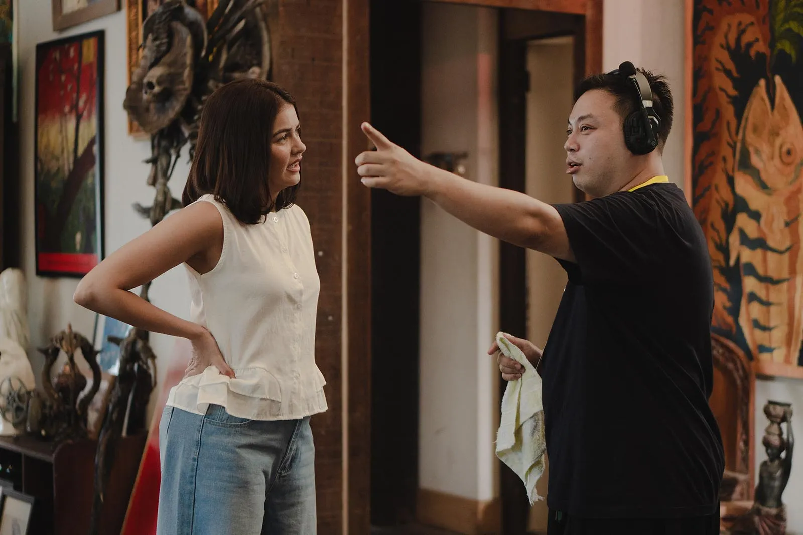 Man with headphones points a finger at a woman in a white sleeveless top inside an art gallery, as they chat.
