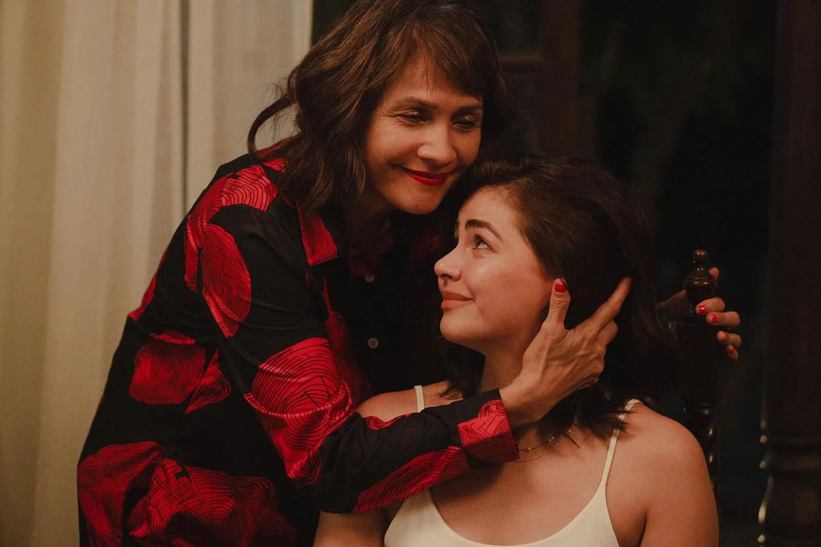 Two women share a tender hug indoors: an older woman with red lipstick cradles the younger woman's face, wearing a red and black patterned jacket and white top.