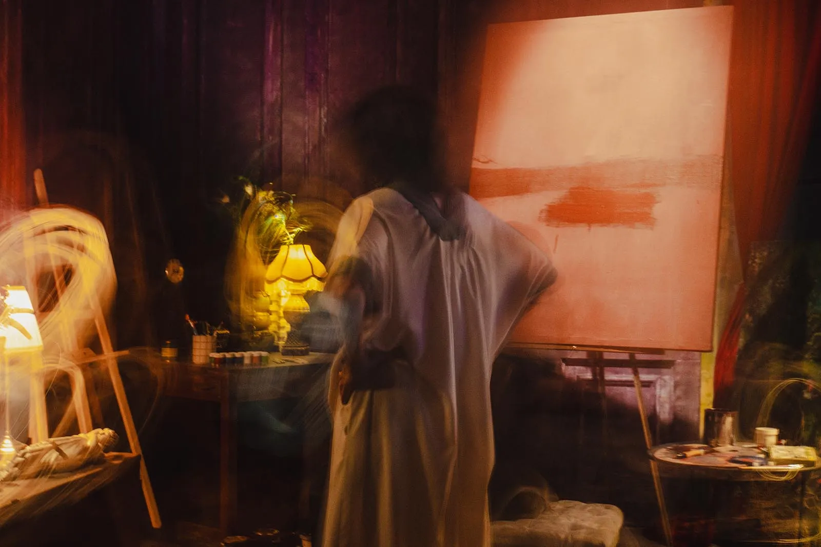 Blurred artist in a dim studio, standing before a large canvas on an easel with warm lamp light.