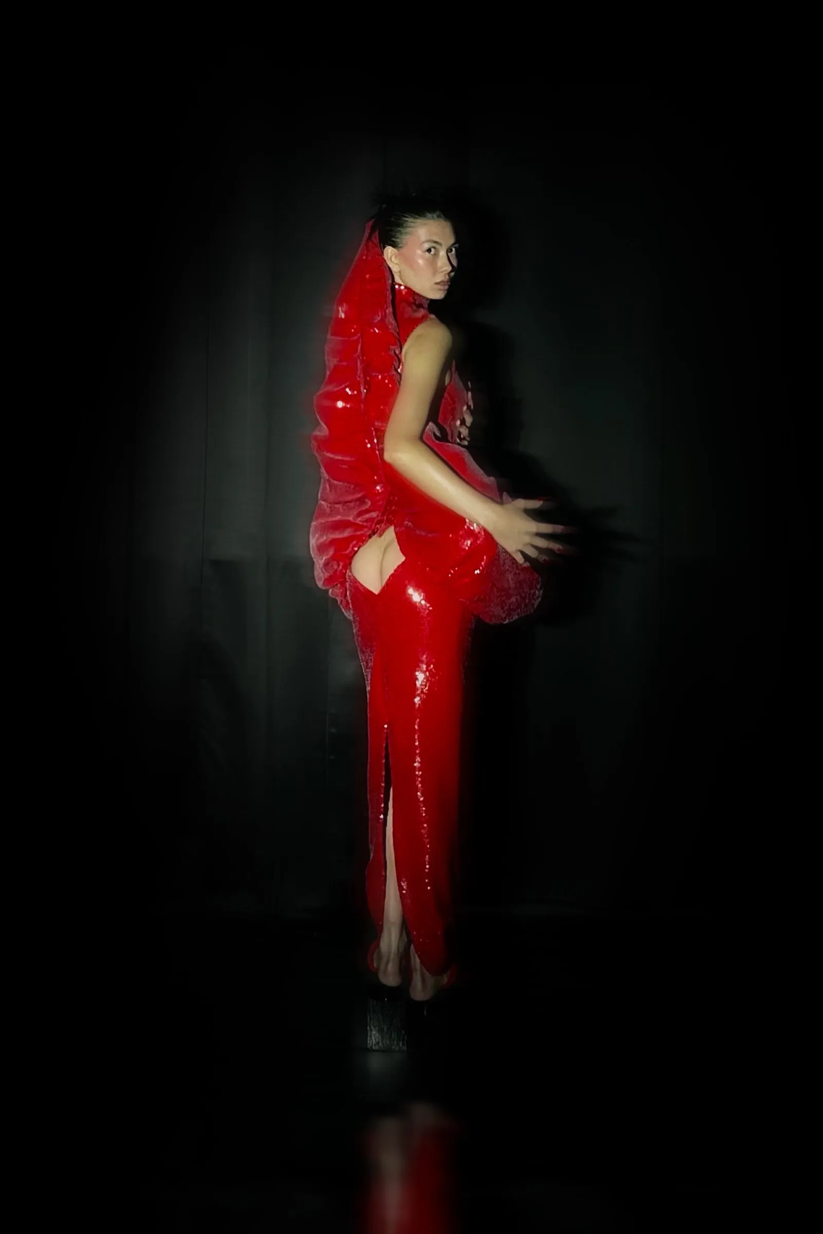 Person in a bright red glossy dress posing against a dark backdrop, dramatic lighting highlighting the outfit.