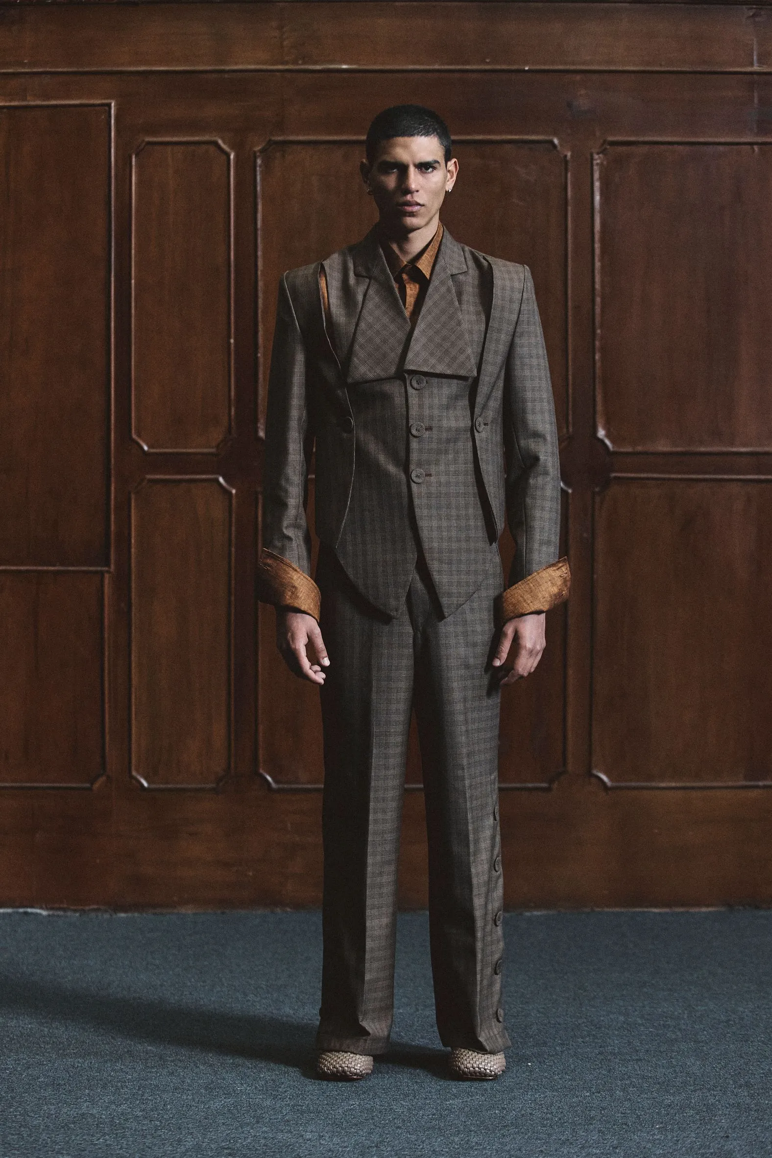 Person standing in a gray plaid three-piece suit against a dark wooden paneled wall, looking forward.
