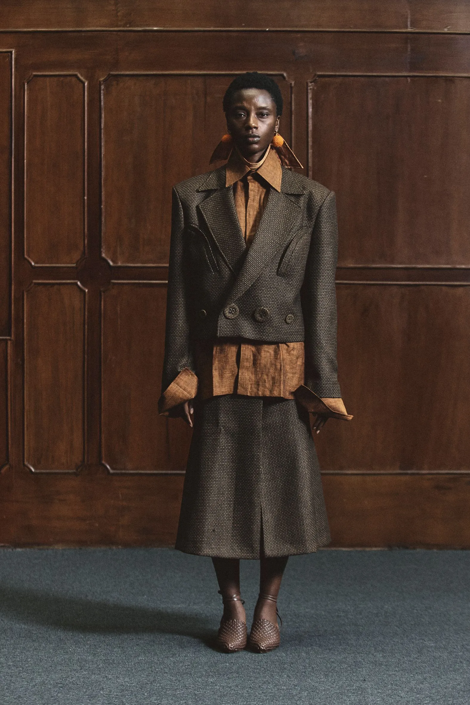 Woman with short natural hair stands against dark wood paneling in a gray tweed double-breasted suit with a brown shirt and oversized collar.