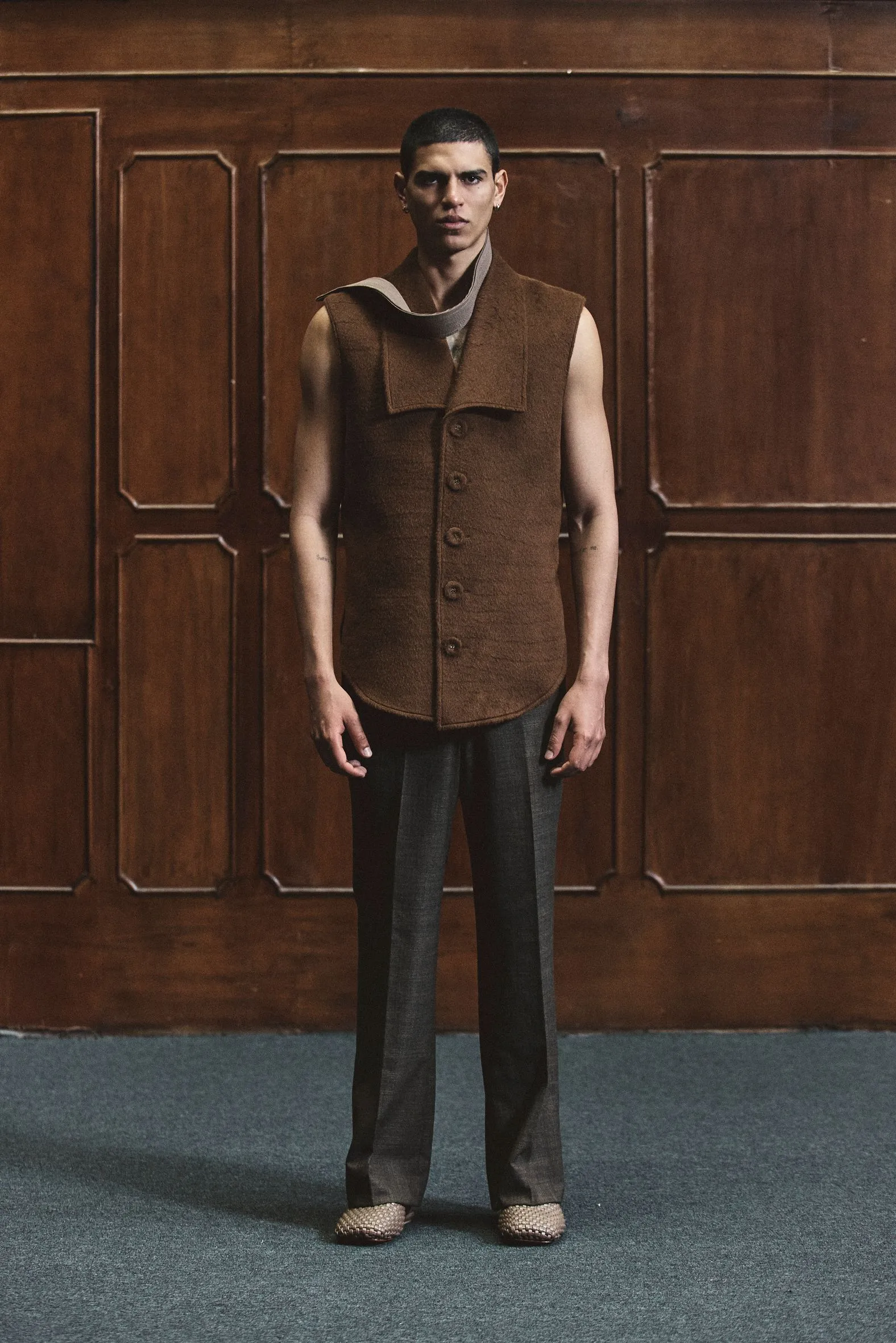 Person standing indoors against dark wooden paneling, wearing a sleeveless brown vest with a loose scarf and dark trousers.