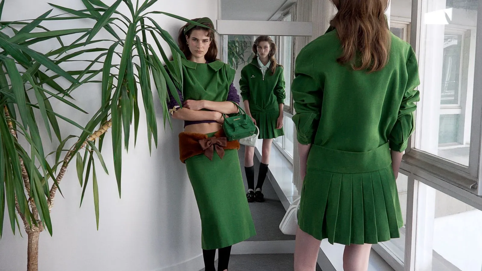 Three models in green outfits pose in a bright corridor with a tall indoor plant; foreground model wears a green skirt and cropped top with a brown fur belt and green handbag.