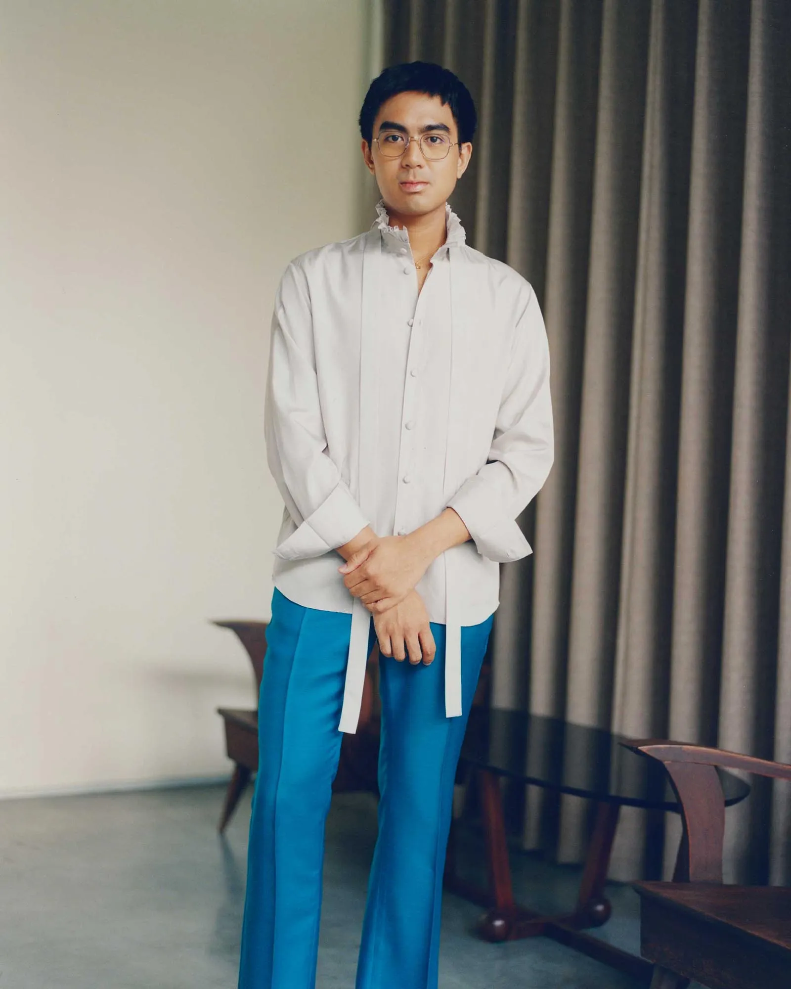 Young man standing indoors in a white kurta with a ruffled collar and blue pants, hands folded in front, against a beige wall and dark curtains.
