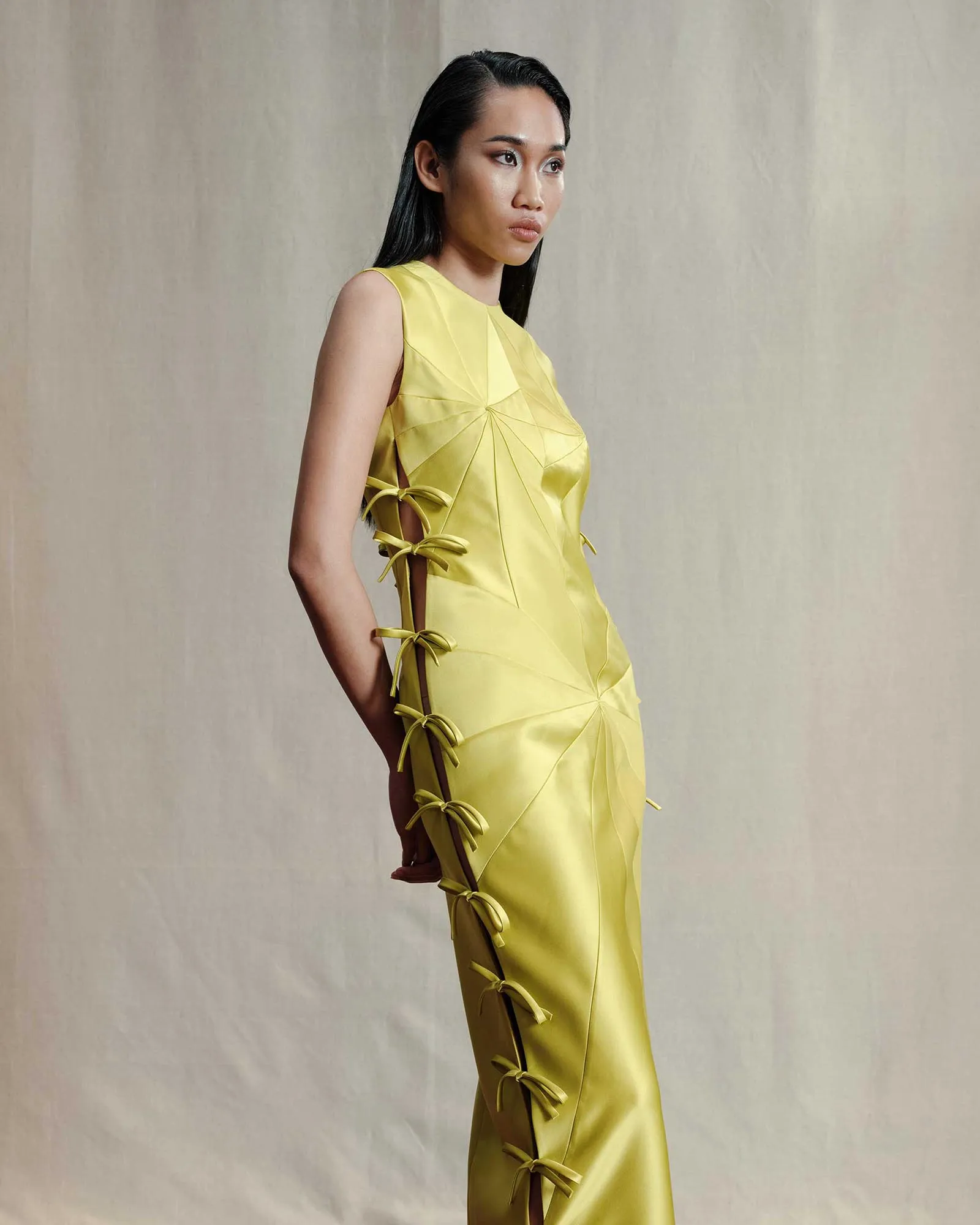 Woman in a sleeveless yellow dress with side lattice-style lacing, posed in profile against a neutral backdrop.