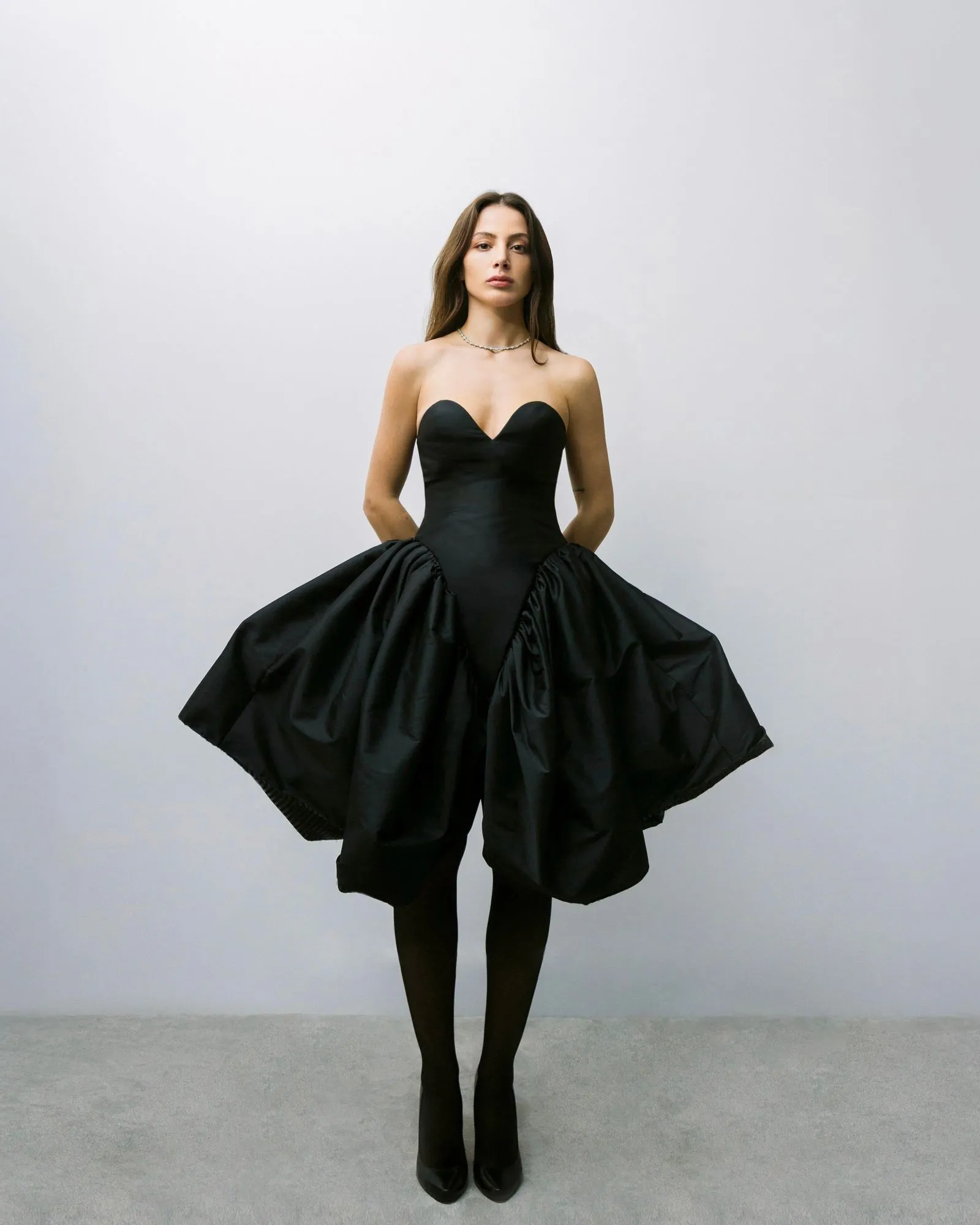 Full-length image of a woman wearing a black strapless ball gown, standing with hands behind her, against a pale gray background.