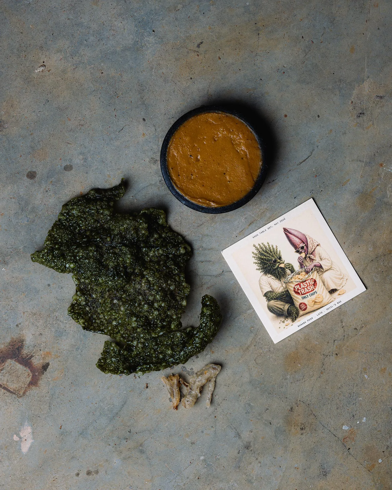 Overhead shot of a dark round dish with orange sauce on a worn concrete surface, beside green kale chips and a magazine with a vintage illustration