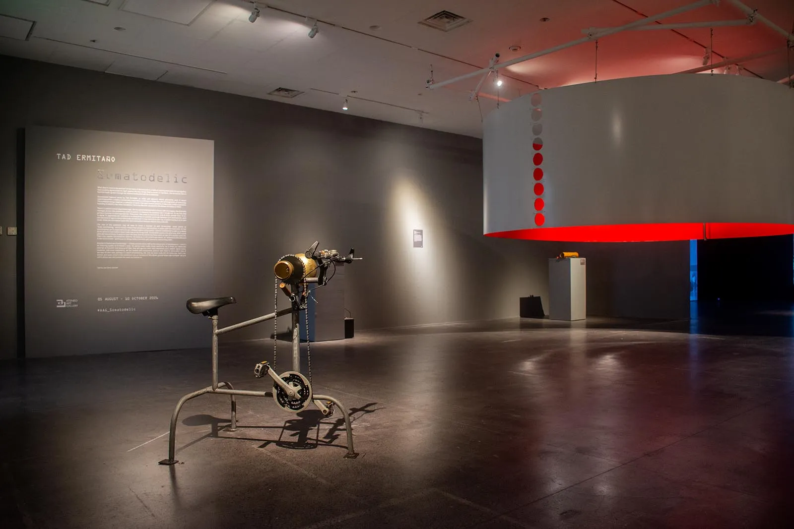 Industrial bike sculpture on a dark gallery floor, with a large white curved installation lit red on the right and a wall plaque in the background.