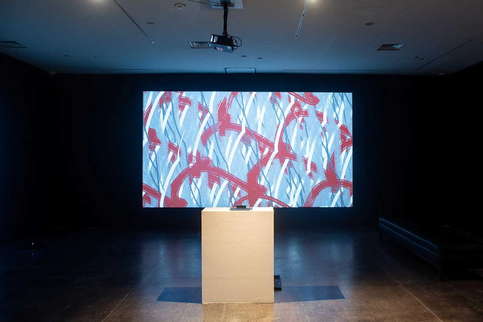 Dim gallery space with a large illuminated abstract artwork on the back wall, featuring blue and red brushstroke patterns, in front of a white pedestal.