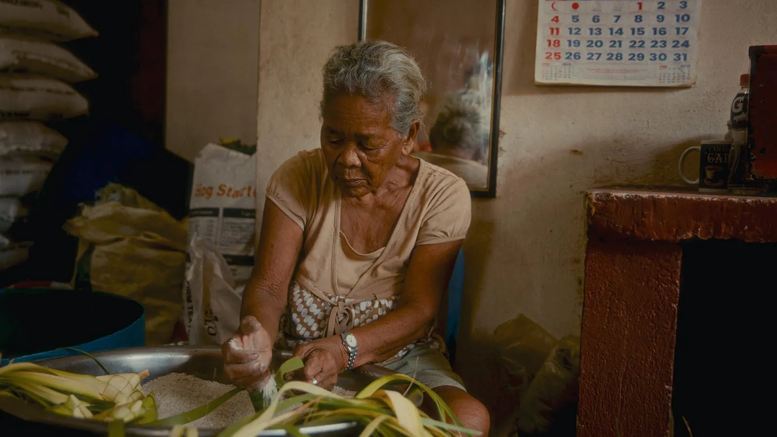 Older woman in a beige top and apron sits at a kitchen table, handling plant leaves over a large metal bowl of grains.