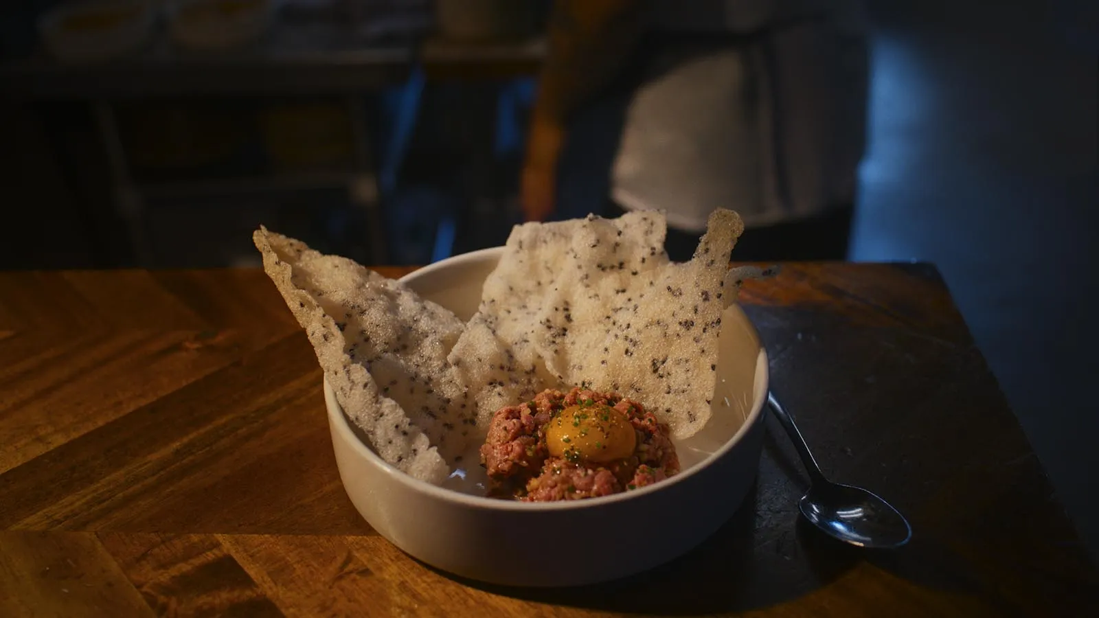 Beef tartare topped with a quail egg, served with crispy crackers in a white bowl on a wooden table.