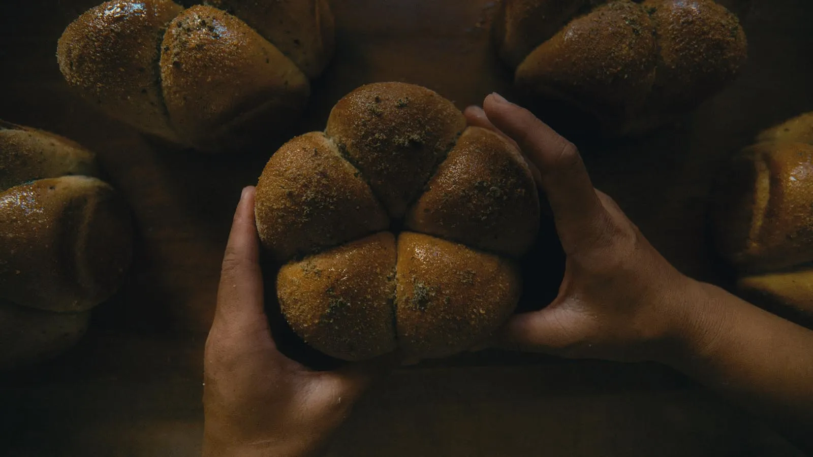 Two hands hold a cluster of six golden-brown, sesame-seed–topped dinner rolls pressed together in a single loaf.