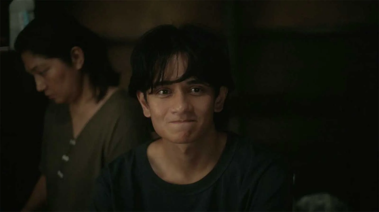 Young man in the foreground with dark hair and a subtle smile, in a dimly lit room; another person sits in the background looking down