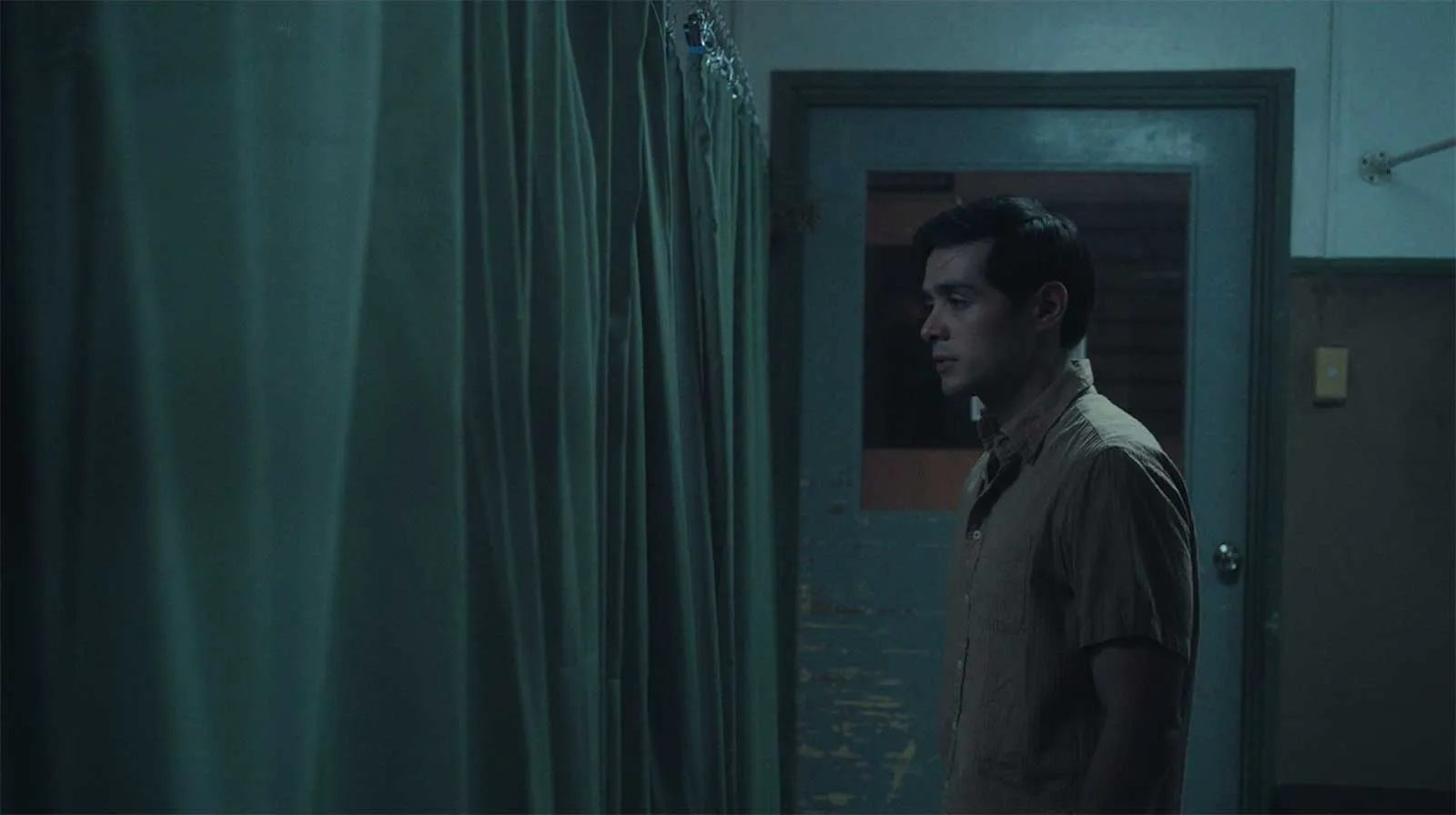 Man in a beige shirt stands in a dim teal-lit hallway, beside hanging curtains and a closed door, looking to the right.