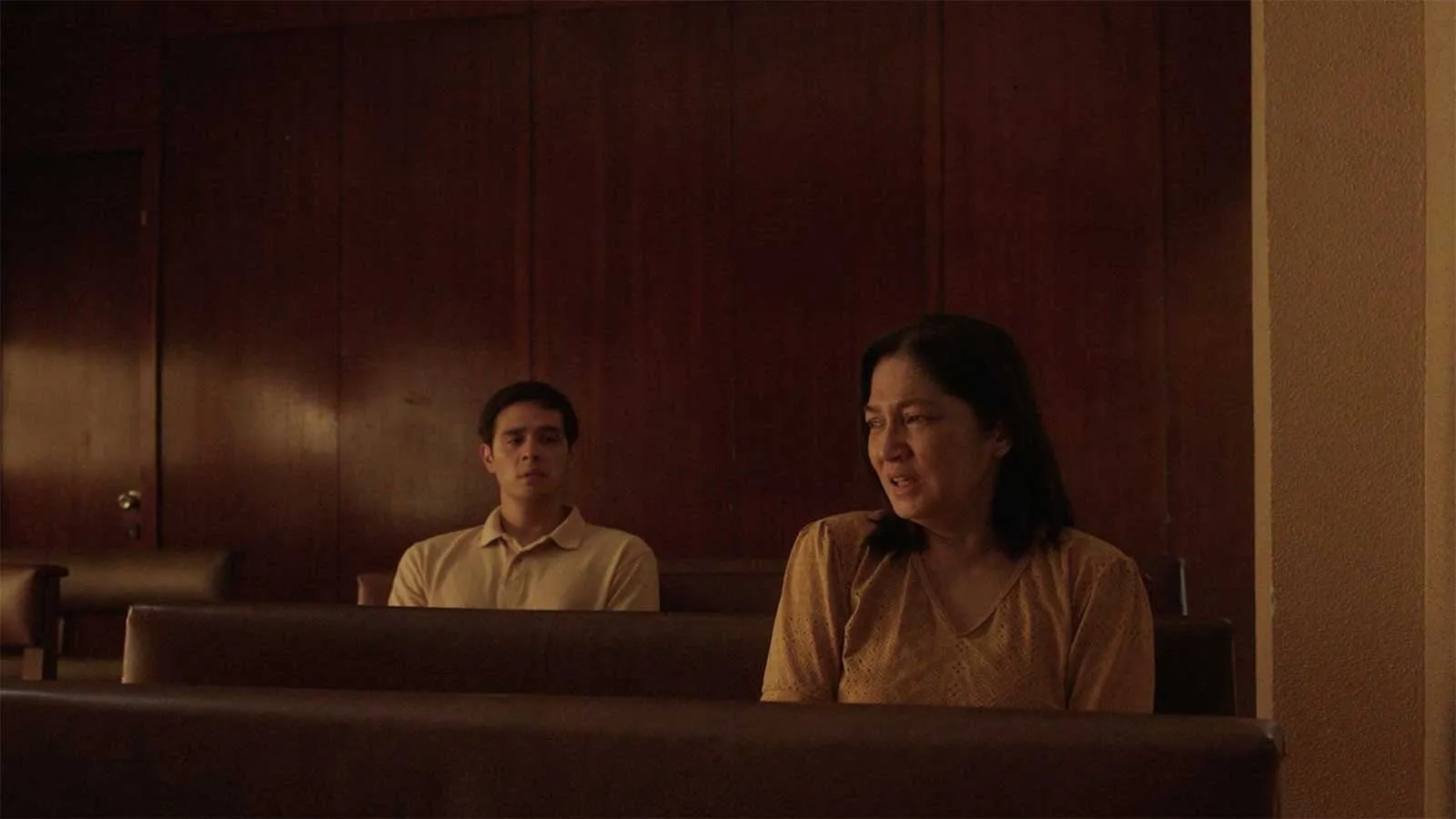 Two people sit apart on wooden benches in a dim room, one man in a light polo and a woman in a peach blouse looks off to the side.