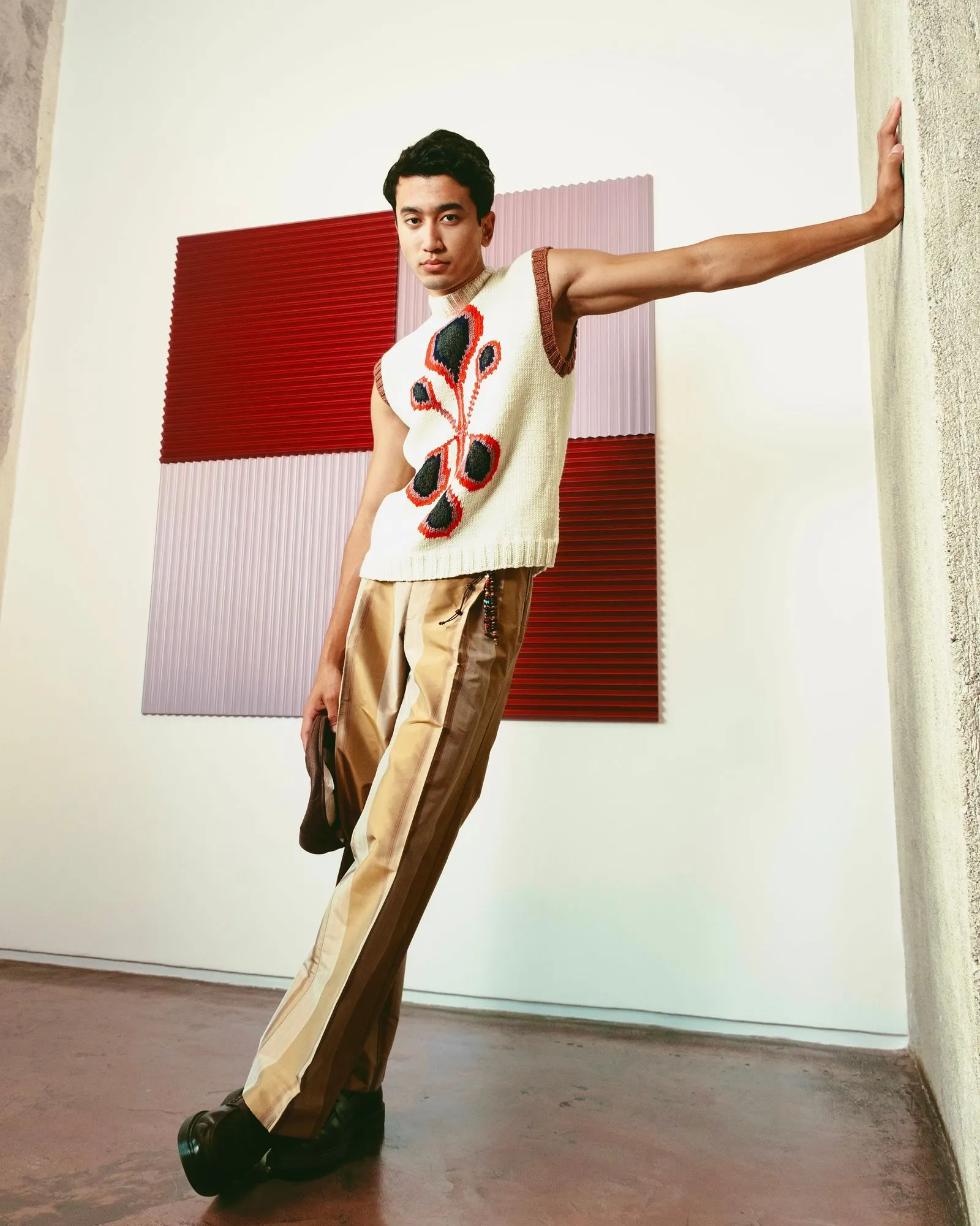 Young man in a sleeveless knit sweater with a red and black butterfly design leans against a textured wall, arm extended to the side with bold fashion-forward pose.