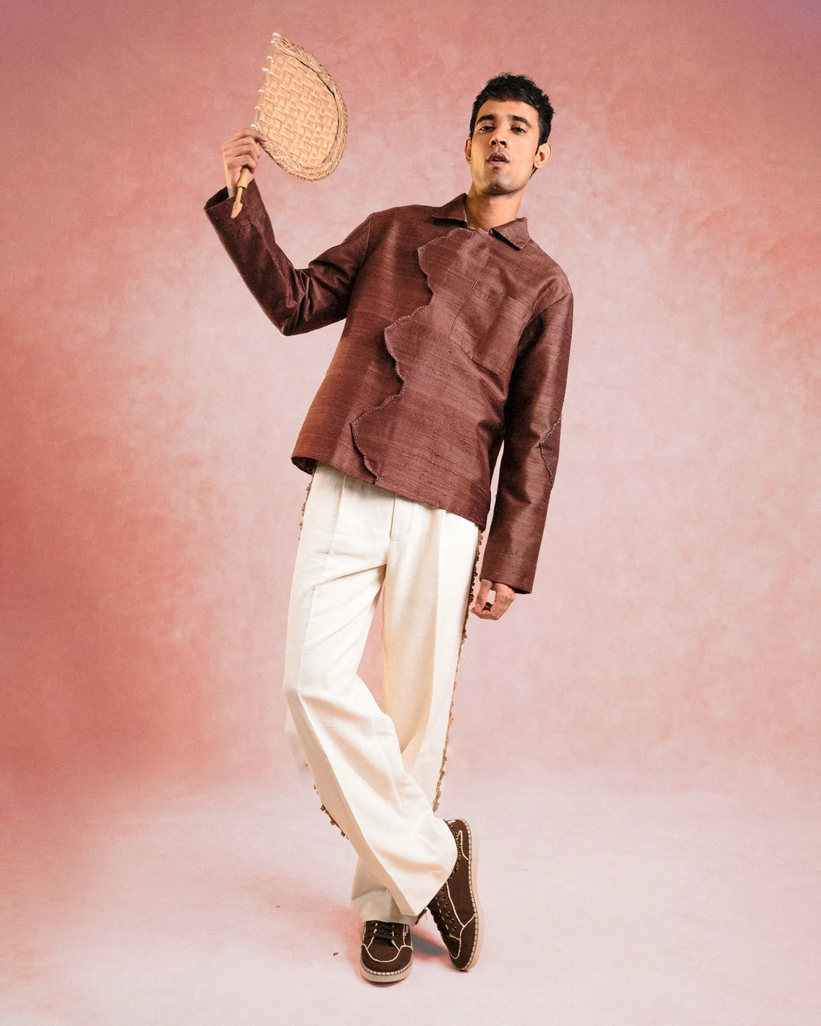 Man in a brown textured shirt and light trousers stands against a pink backdrop, holding a woven wicker fan aloft in his right hand as he poses.