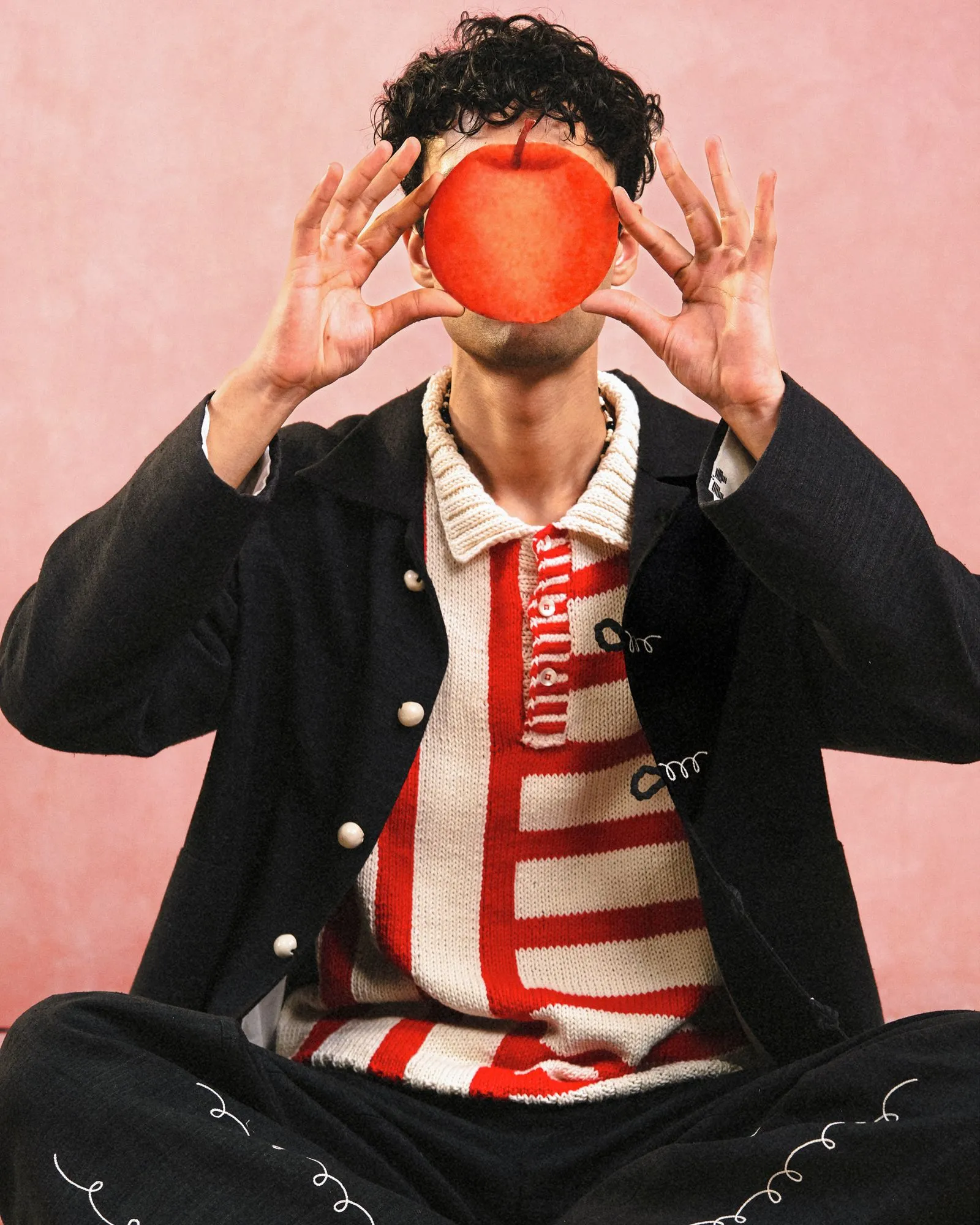 Person holding a red apple in front of their face, obscuring features, against a pink background.